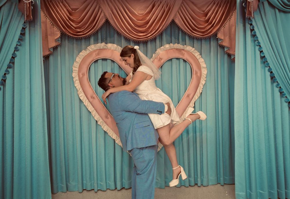 A man in a light blue suit lifts a smiling bride in a white dress in front of a heart-shaped frame and teal curtains.