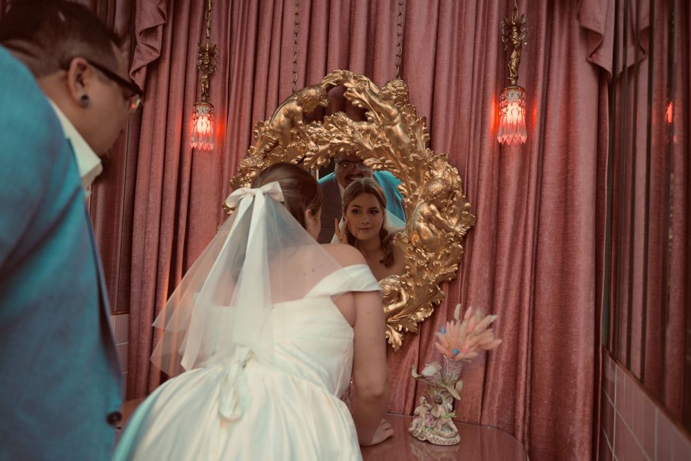 Bride in a white gown and veil gazes into an ornate gold mirror beside a man in a teal jacket, with pink curtains and warm lanterns behind.