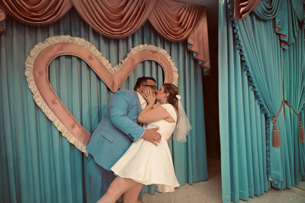 A bride in a short white dress and a groom in a blue suit kiss in front of a heart-shaped frame and teal curtains at a wedding chapel.