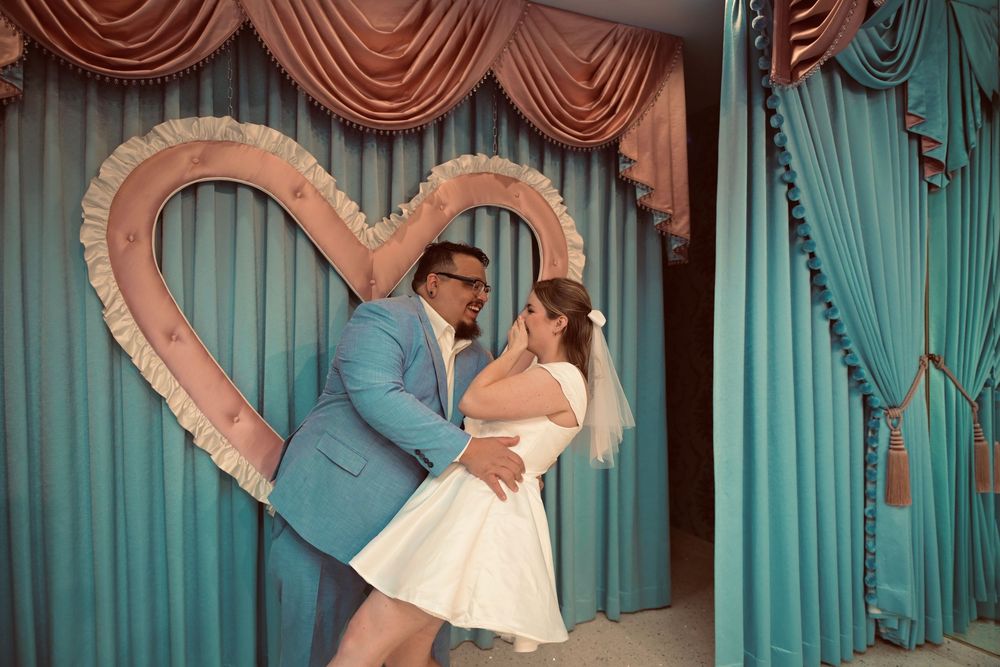 Couple in a blue suit and white dress embrace in front of a heart-shaped frame and teal curtains at a wedding chapel.