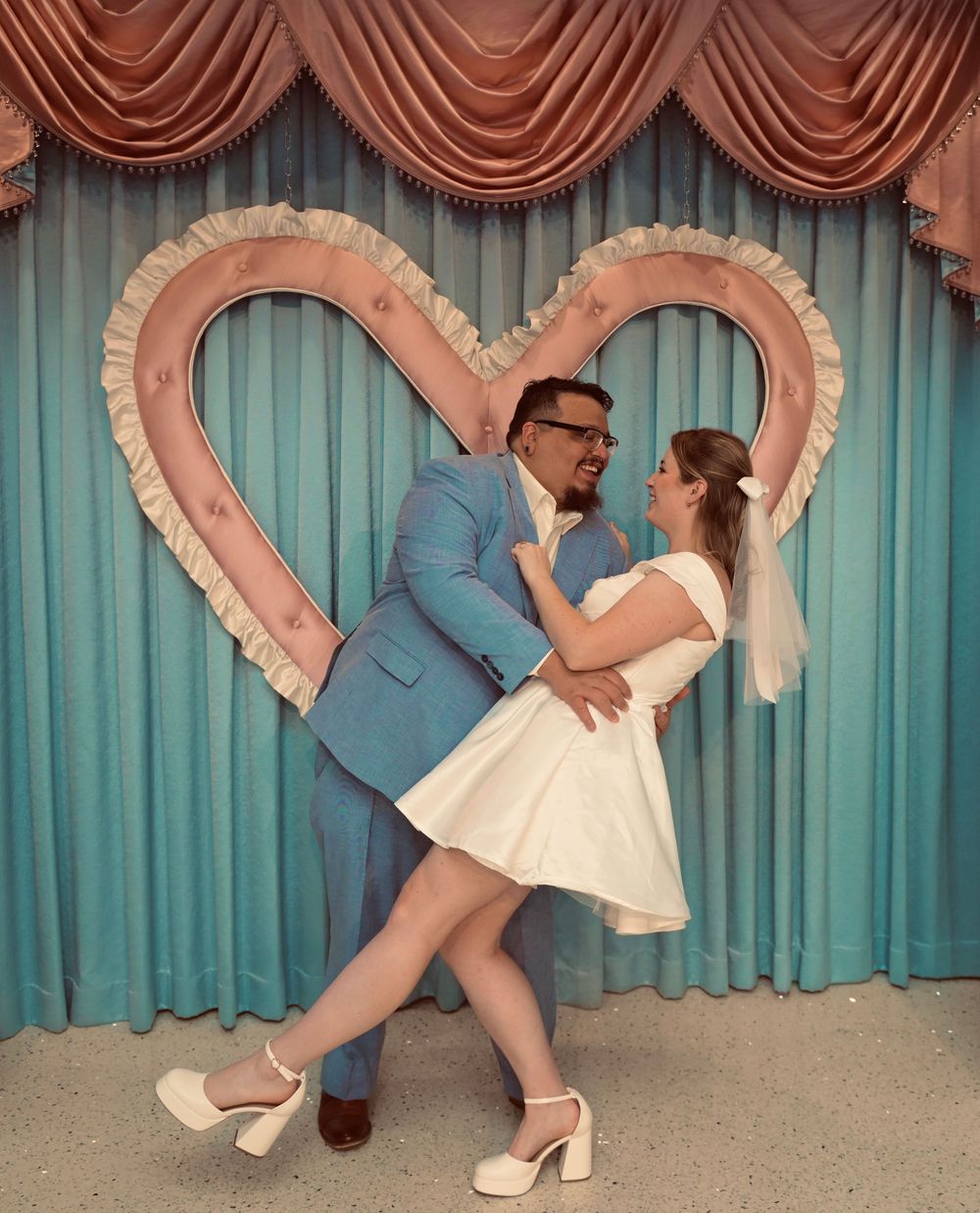 A bride in a short white dress and veil is dipped by her groom in a blue suit before a heart-shaped backdrop and teal curtains.