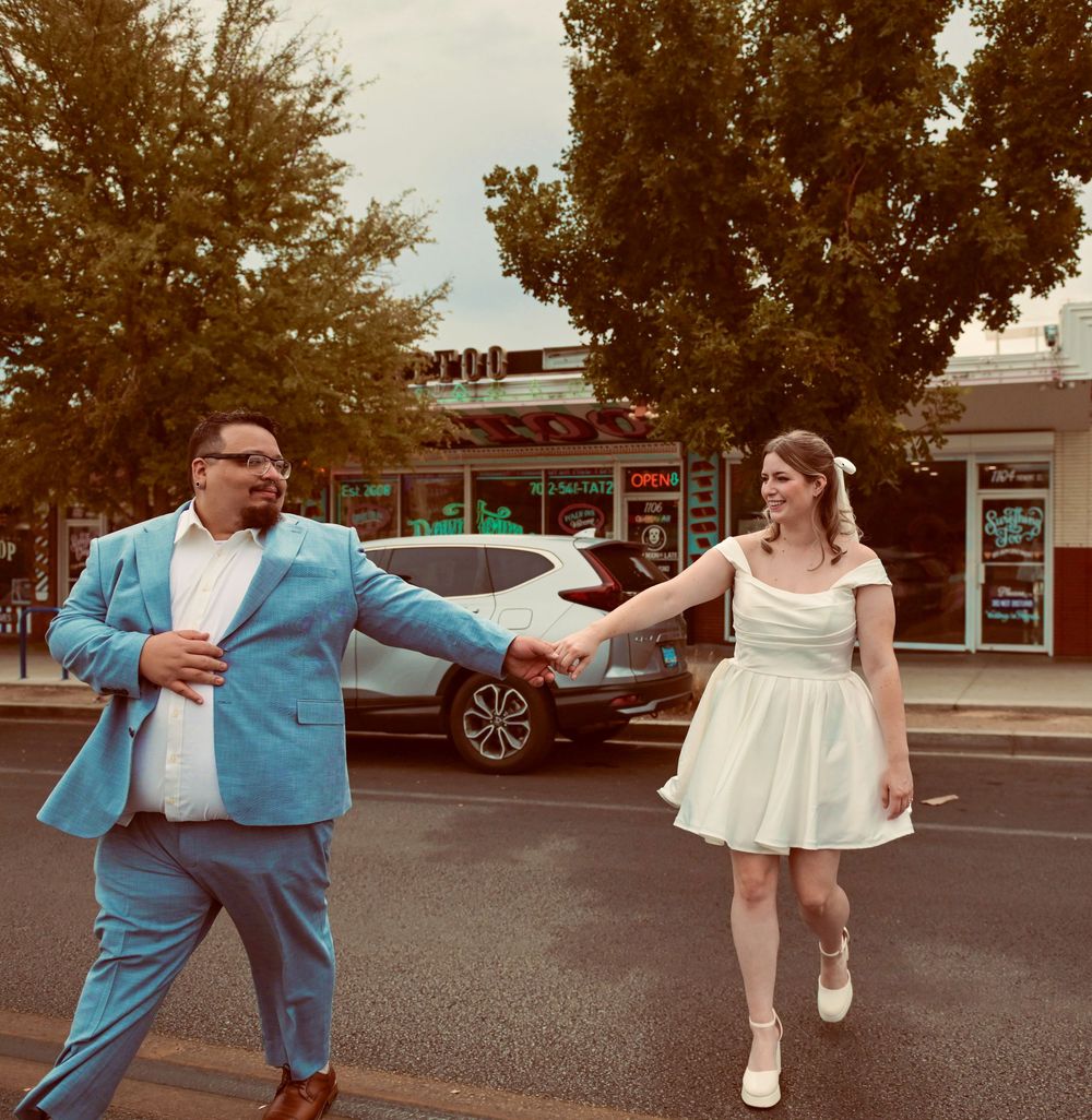 A groom in a turquoise suit holds hands with his bride in a short white dress as they walk across a city street.