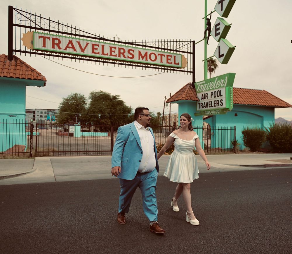 Bride in a white dress and groom in a turquoise suit walk across the street in front of the vintage Travelers Motel sign.