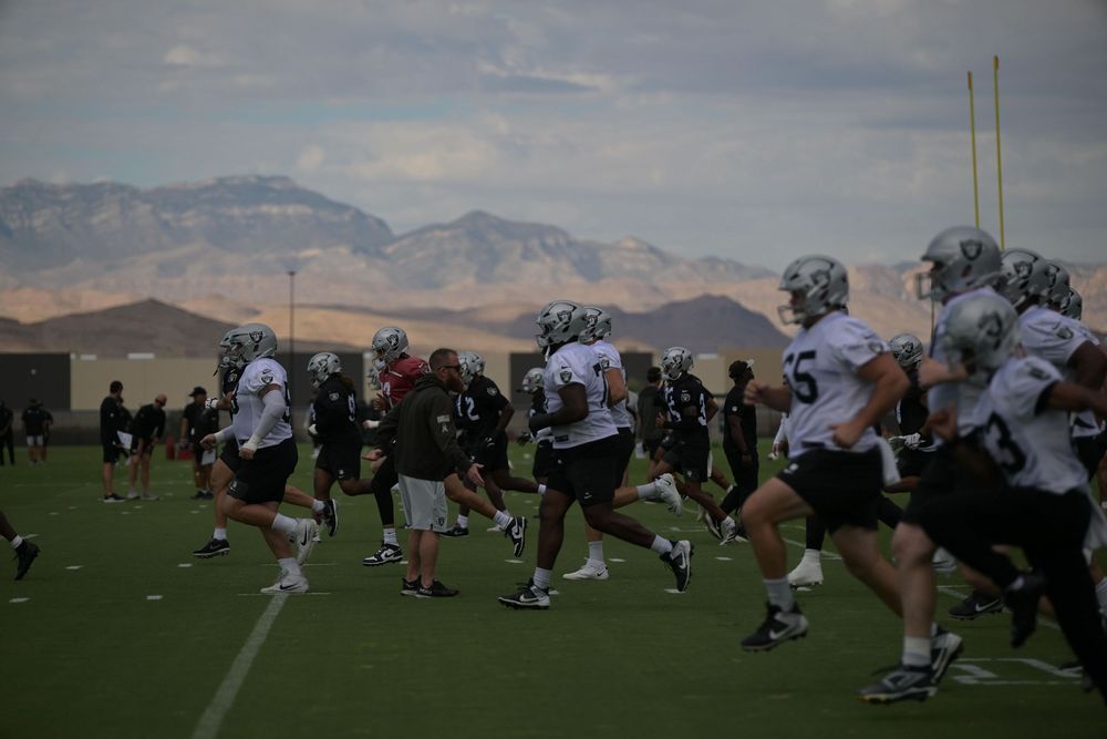 Football players in white and black practice jerseys run drills on a green field with desert mountains and a hazy sky in the background.