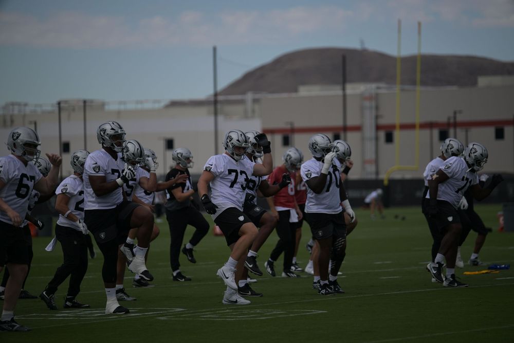 Football players in white jerseys and helmets stretch and warm up together on a practice field with a hill and stadium in the background.