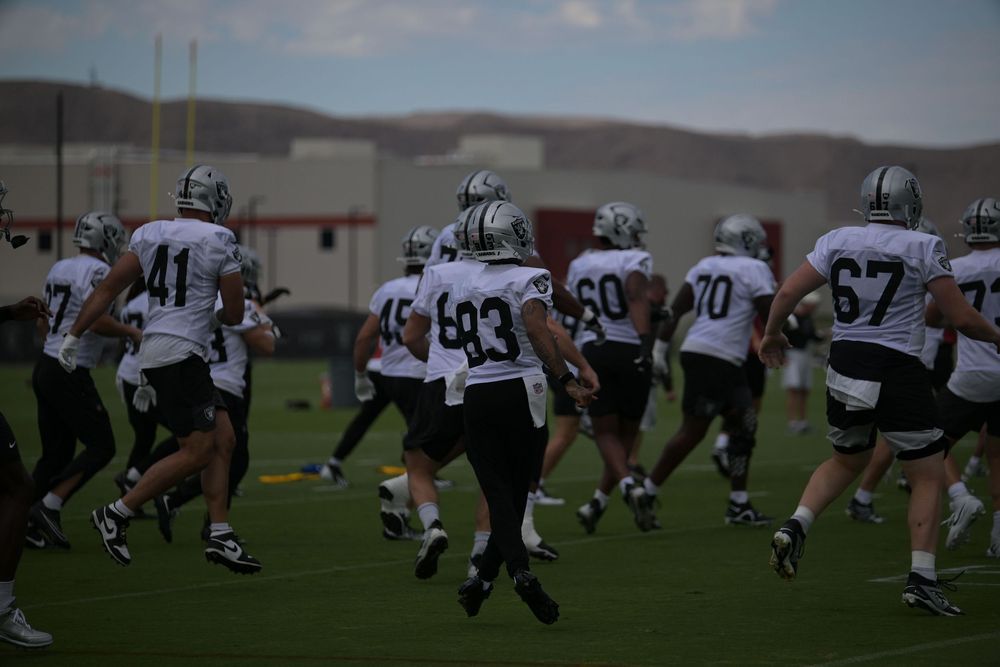 Football players in white jerseys and helmets jog across a practice field during a team workout with hills in the background.