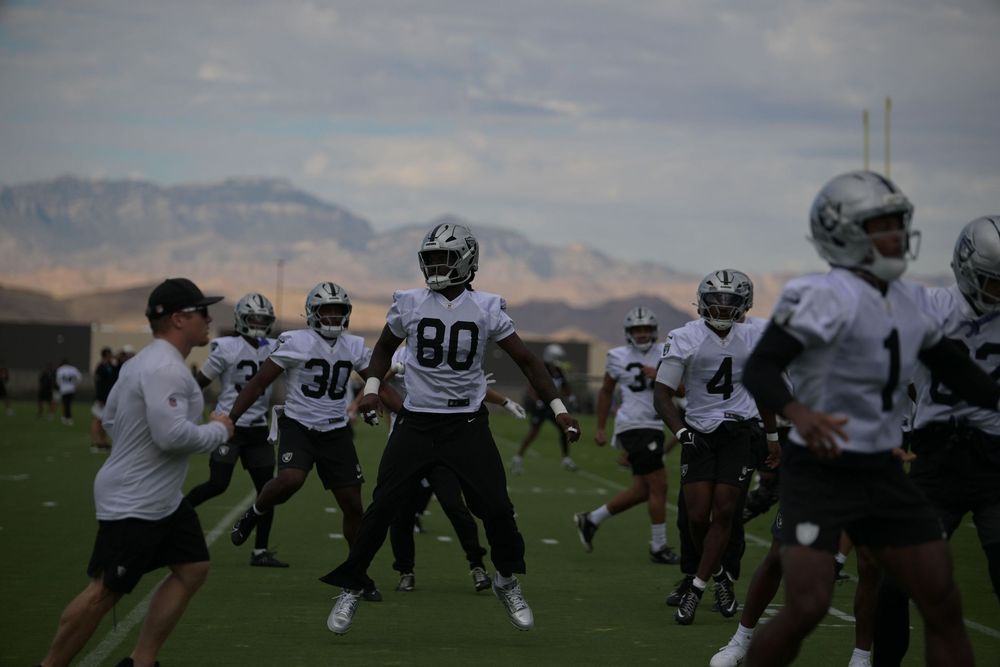 Football players in white jerseys and helmets run drills on a practice field while a coach watches, with desert mountains in the background.