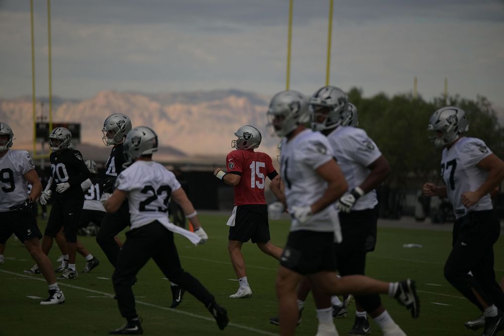 Football players in white jerseys and helmets jog across a practice field with desert mountains in the background.