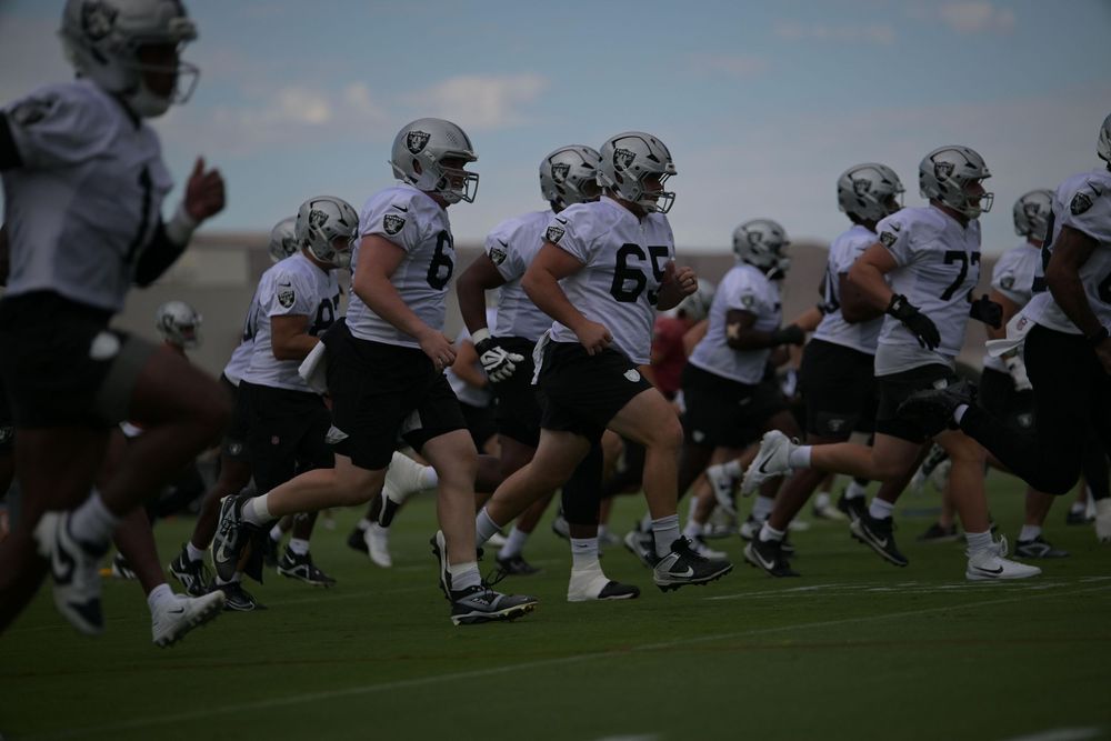 Football players in white jerseys and helmets run together in formation across a grass field during an outdoor team practice session.