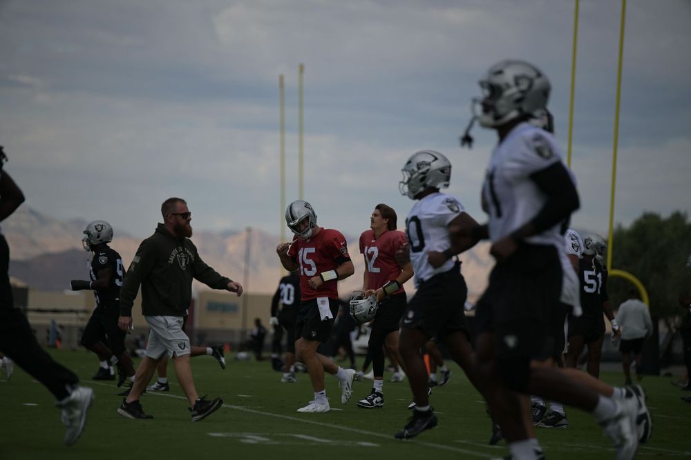 Players in helmets and jerseys jog across a practice field as a coach in a dark jacket watches, with mountains behind them at dusk.