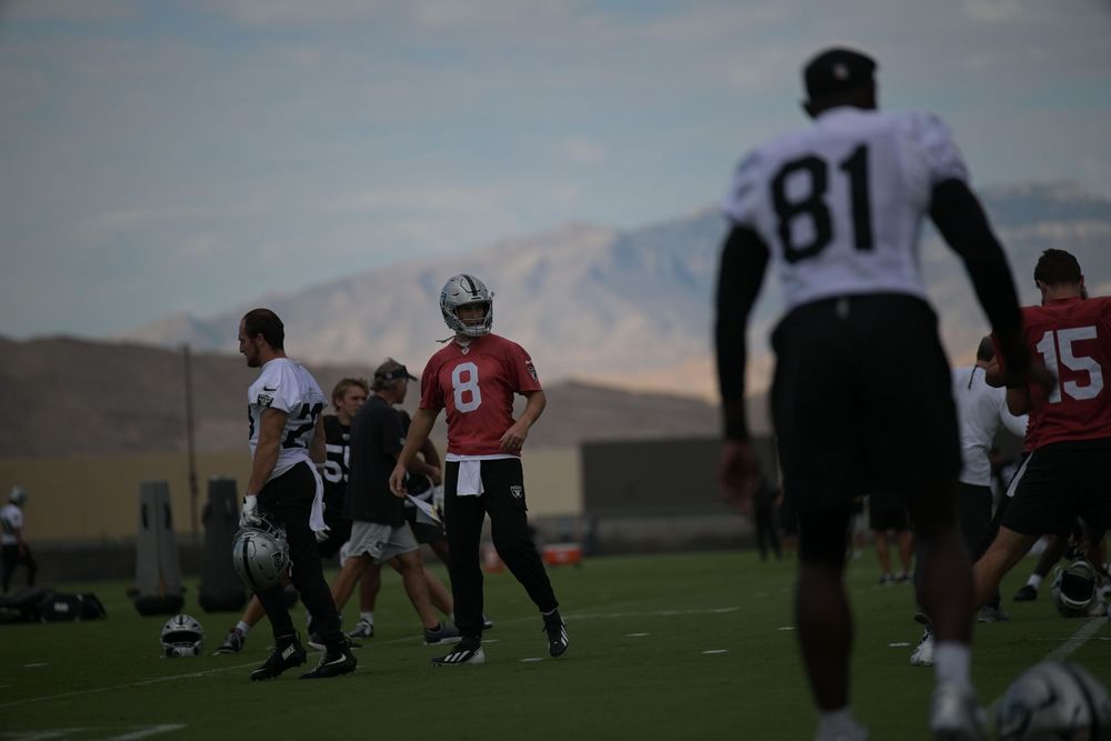 Football players in practice gear run drills on a grass field at training camp, with a quarterback in a red jersey and mountains behind.