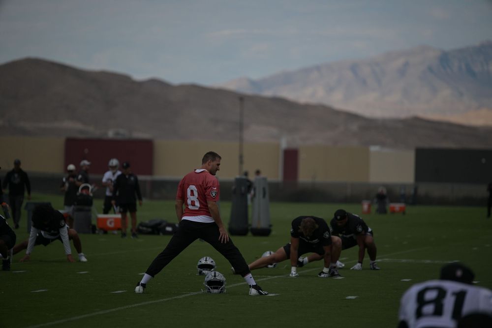 A football player in a red jersey stretches on a practice field while teammates warm up near mountains at dusk.