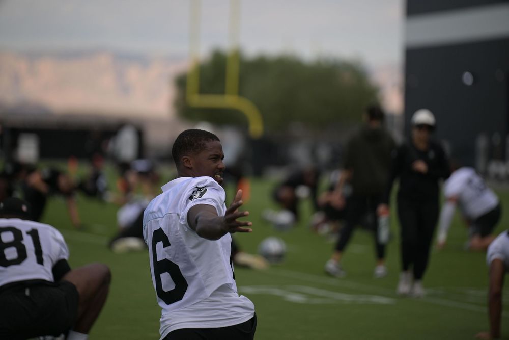 Football player in a white number 6 jersey stretches on a practice field while teammates warm up in the background.