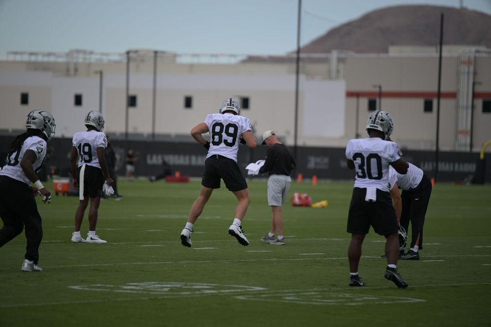 Football players in white jerseys and black pants run drills on a practice field, with numbers 89 and 30 visible and a desert hill behind.