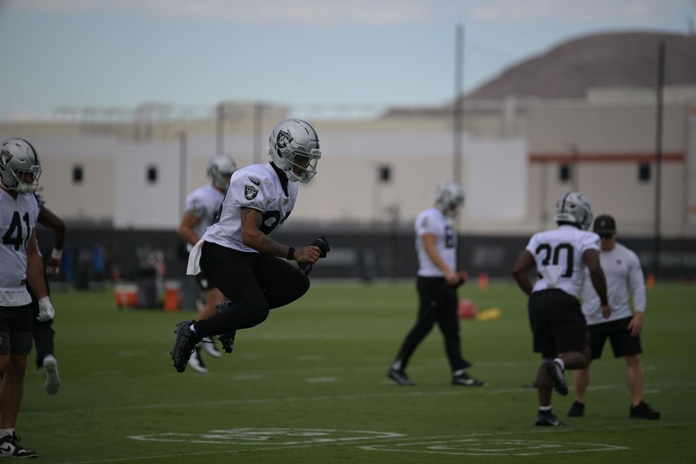 Football players in white practice jerseys and silver helmets run drills on a grass field during an outdoor team practice.