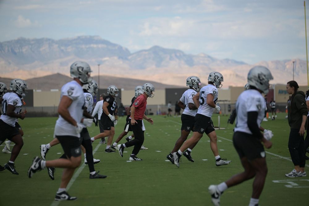 Raiders players in white practice jerseys stretch and run drills on a grass field with a mountain range rising behind the facility.