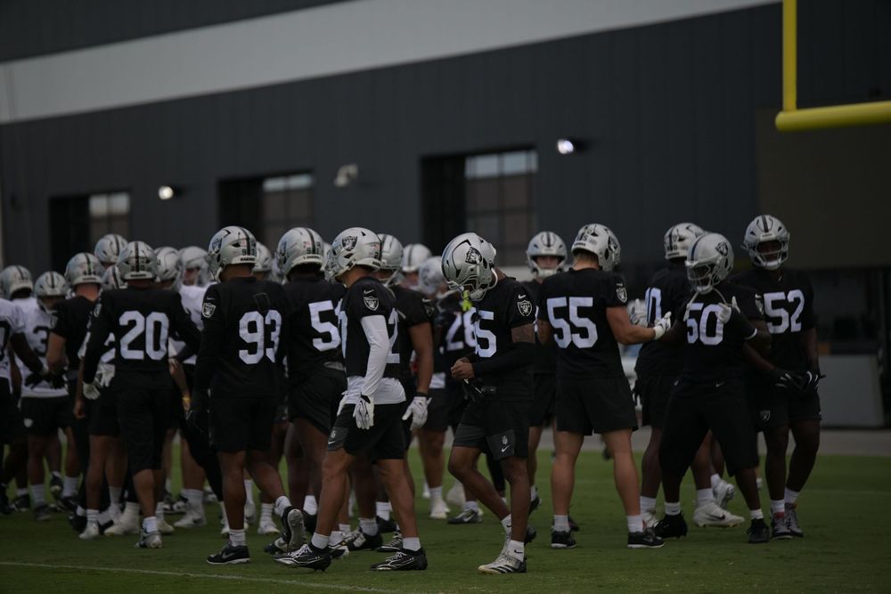 Football players in dark jerseys and silver helmets gather in a group on a practice field during an evening team session.