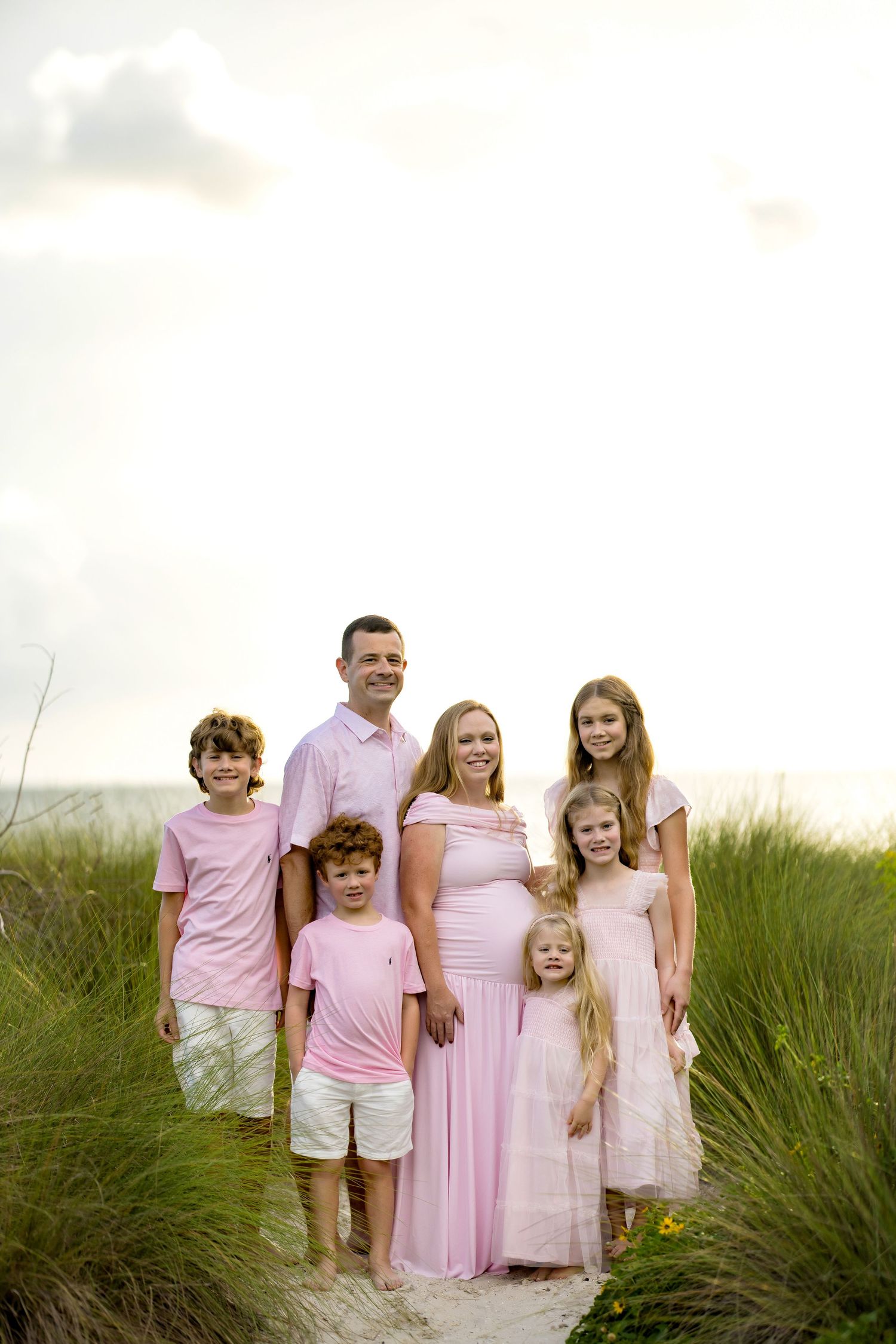 Family of seven in coordinating pink outfits posing on a sandy beach path surrounded by green grass.