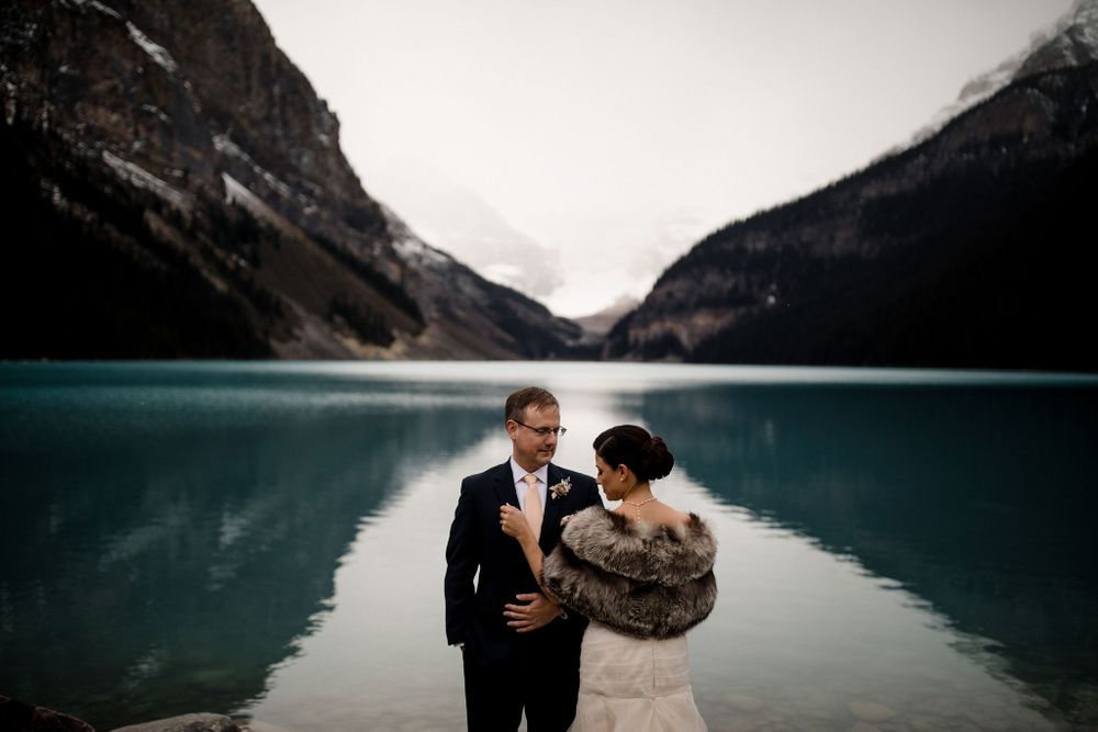 A couple shares a romantic kiss on a rocky mountain overlook with a turquoise lake and dramatic peaks in the background.