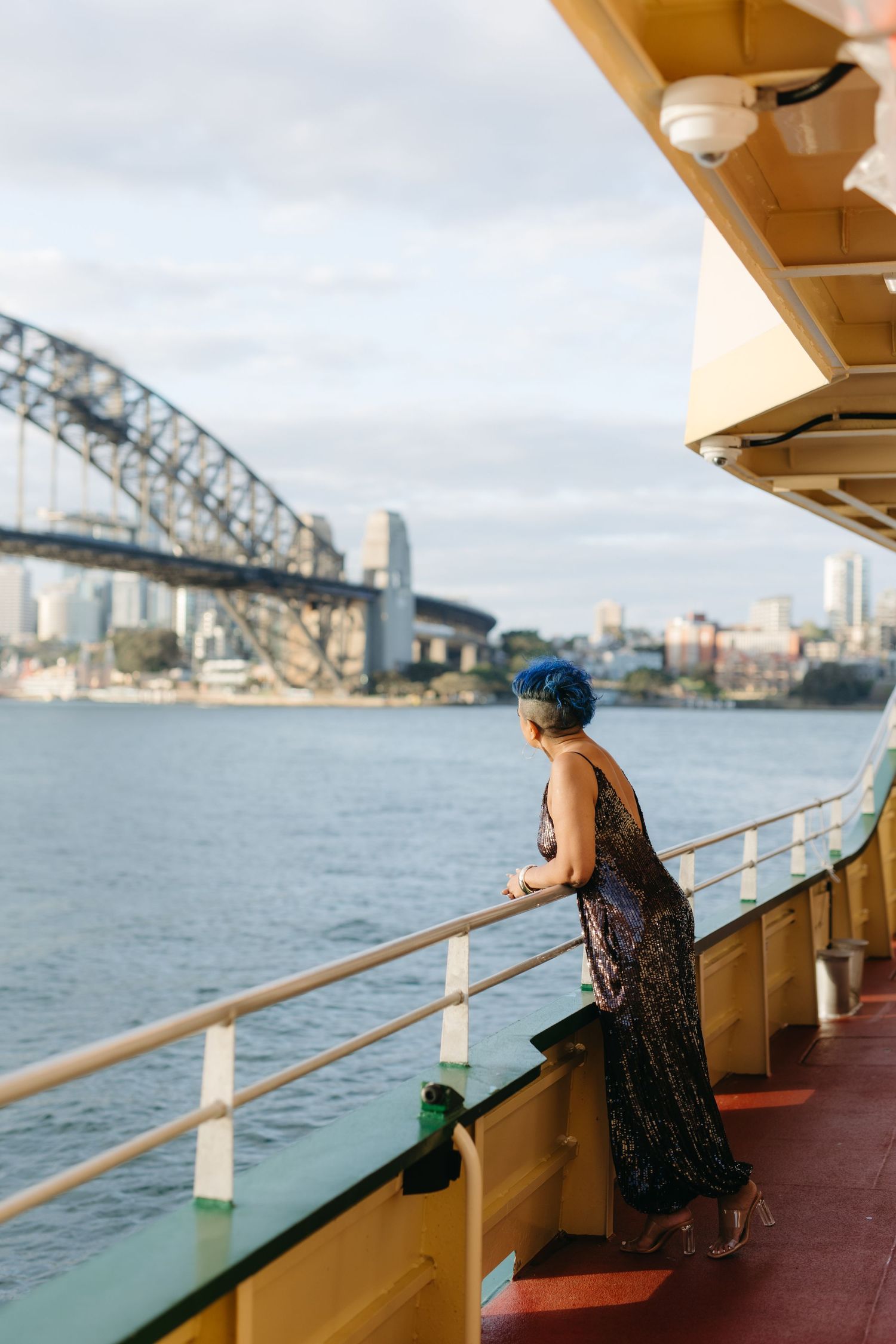 Woman in sequin dress with blue hair gazes at Sydney Harbour Bridge from a ferry deck.