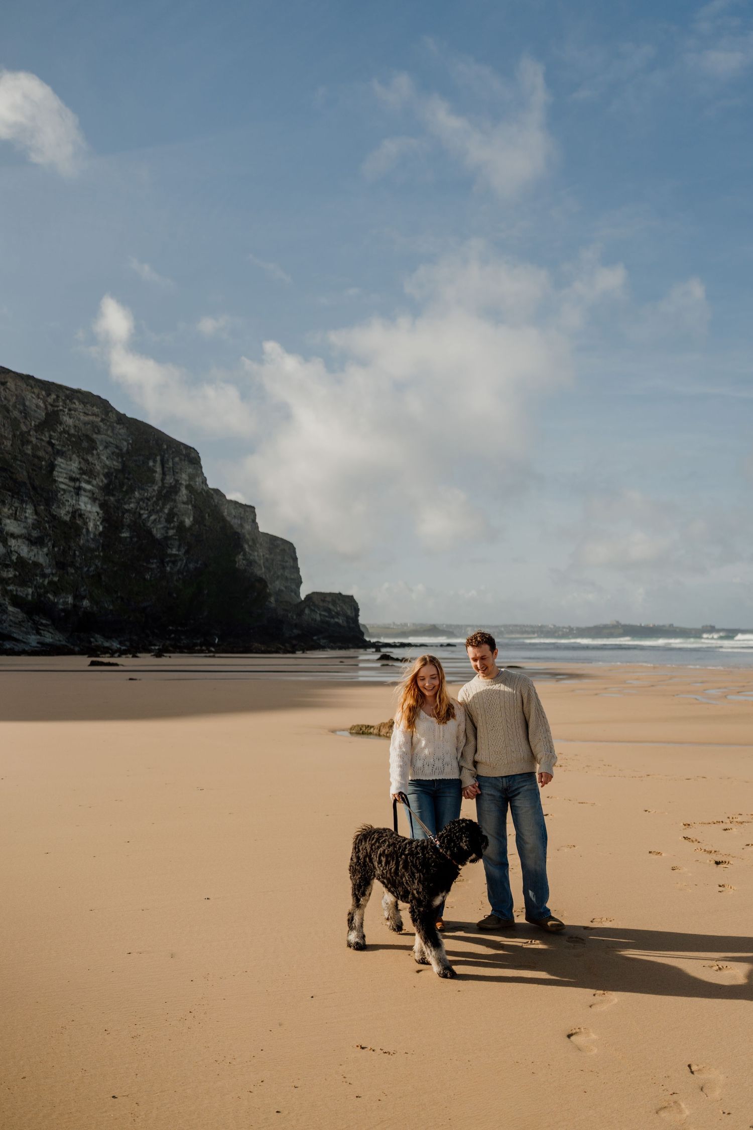 Couple walking a black dog on a sandy beach with dramatic cliffs and blue sky in the background.