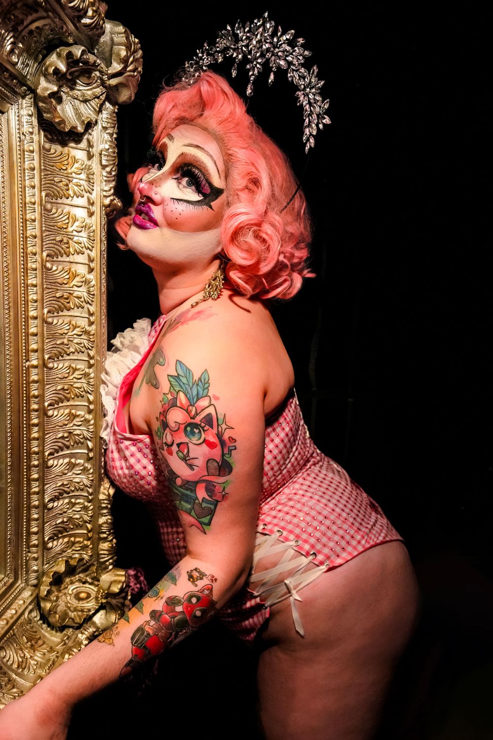 Pin-up style figure in pink gingham swimsuit with red hair, tattoos and crown poses against ornate gold mirror frame.