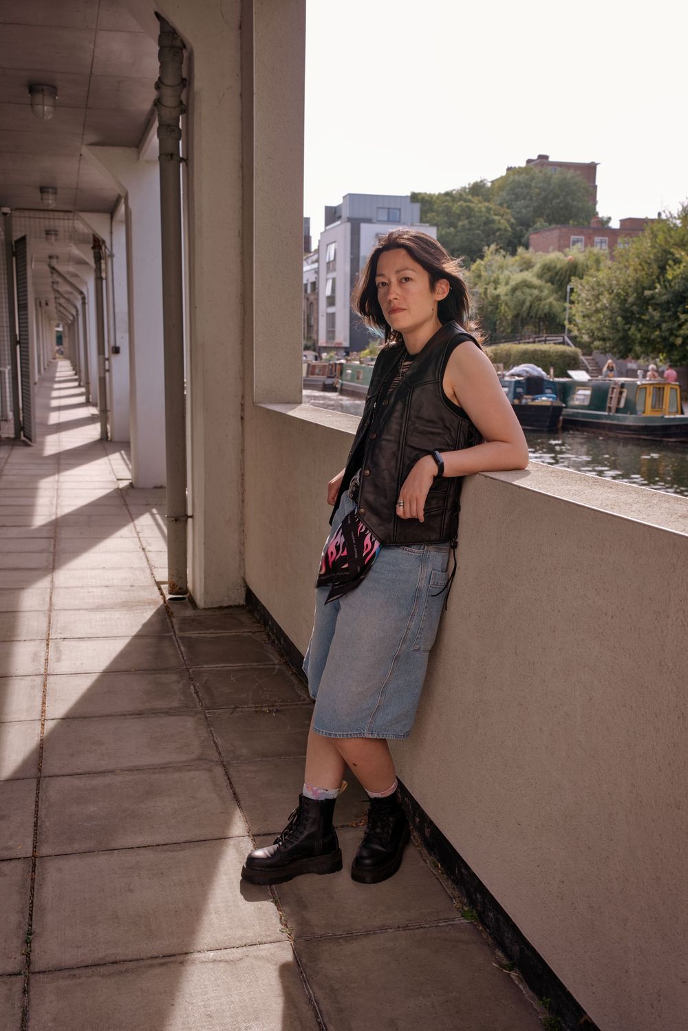 Young woman in black vest and denim skirt leans against a balcony railing in an urban setting with a waterway behind her.