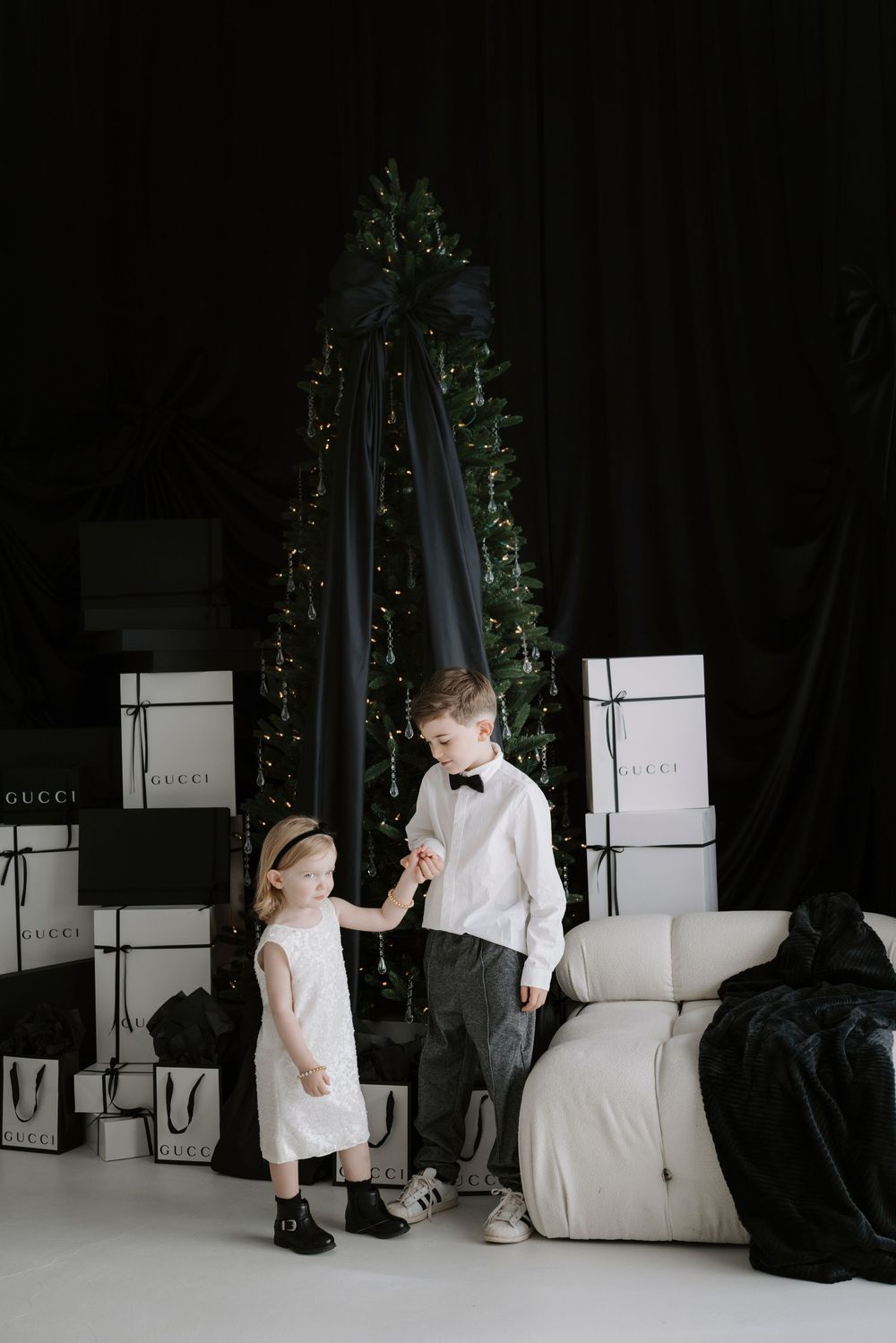 Two elegantly dressed children stand near a Christmas tree surrounded by Gucci gift boxes in a luxury holiday setting.