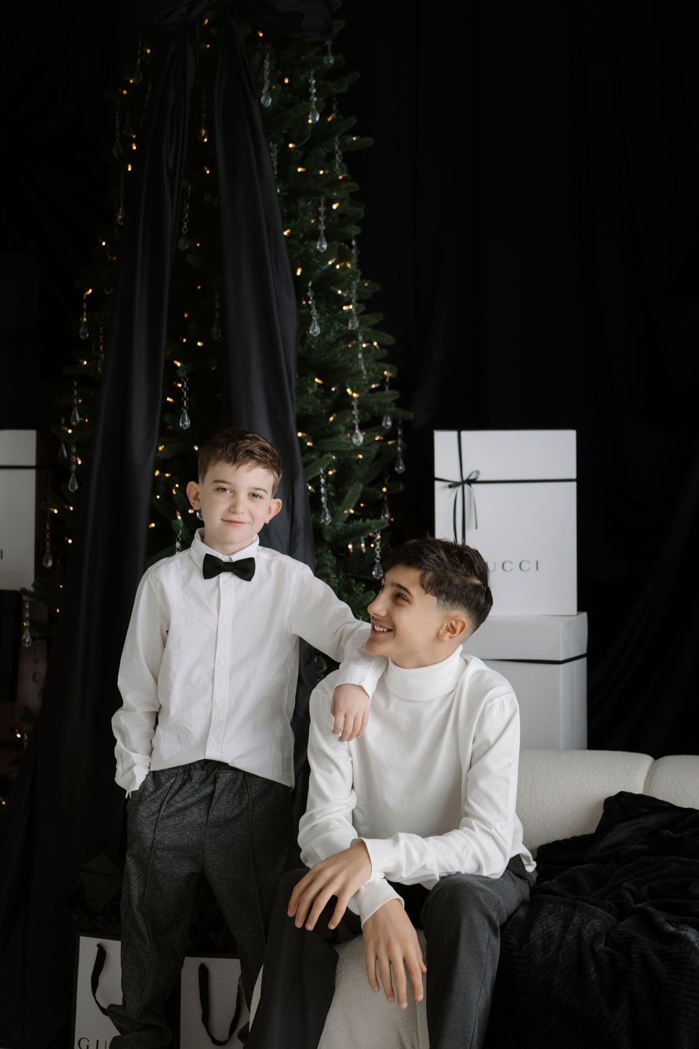 Two boys in white shirts pose near a decorated Christmas tree with Gucci gift boxes in a dark elegant room.
