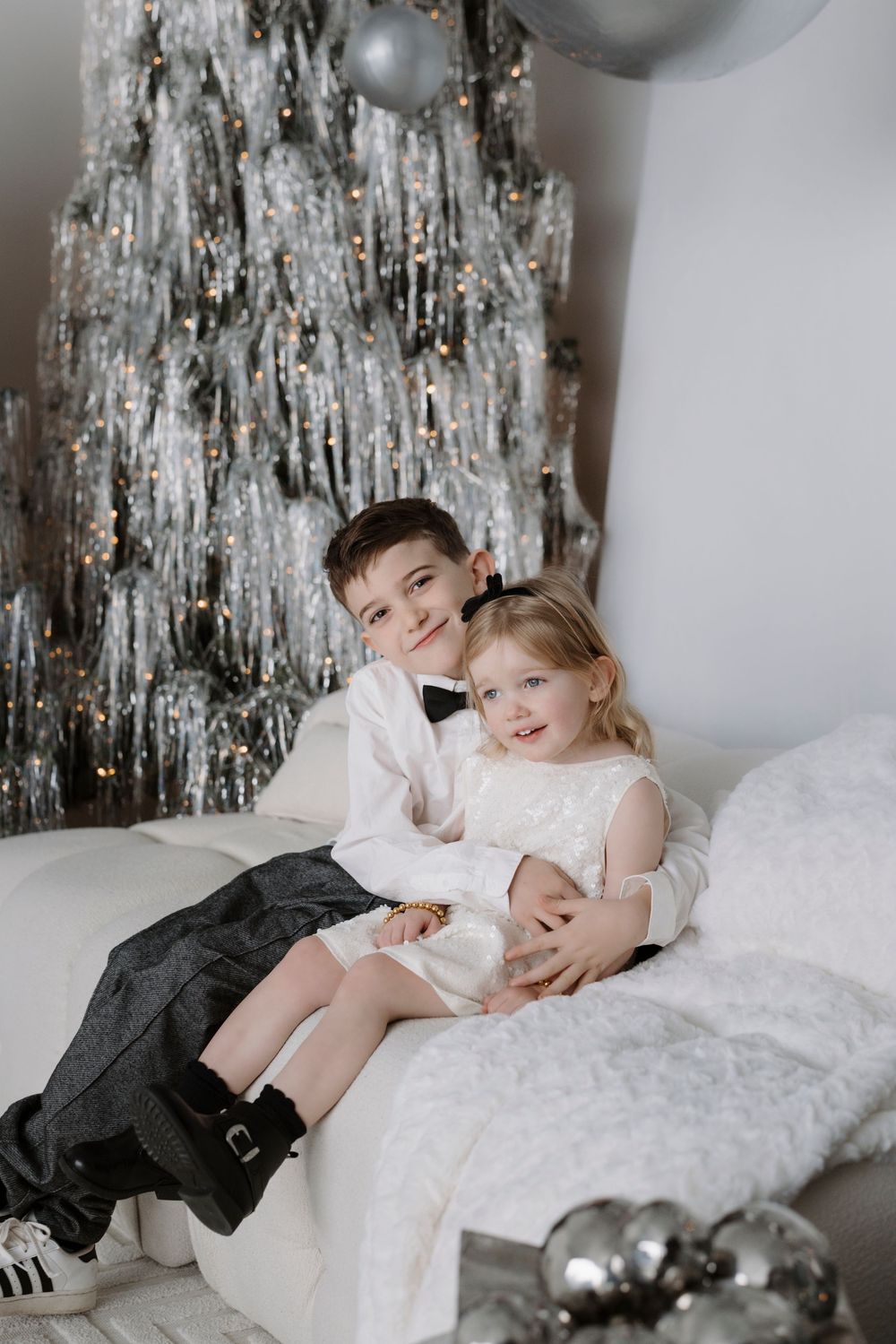 Two children in formal attire sit together on a white sofa in front of a silver Christmas tree.