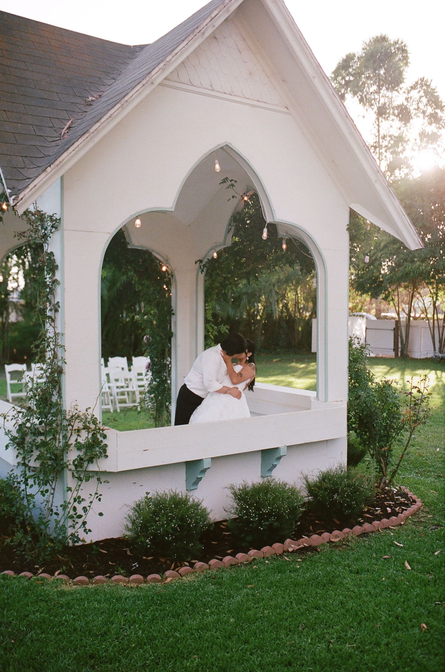 Bride and groom share an intimate embrace inside a white gabled garden chapel at sunset, with string lights and lush greenery.