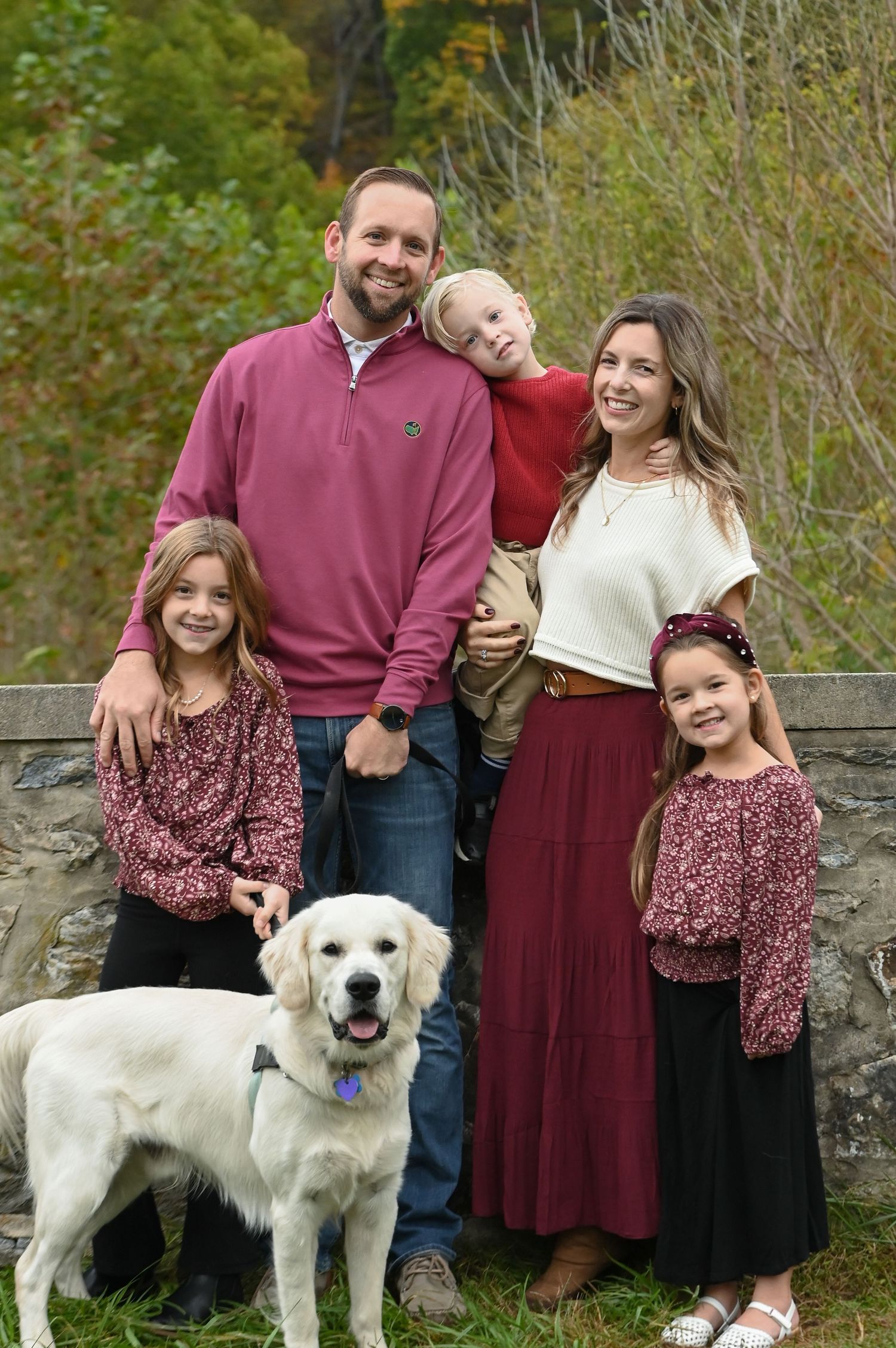 Smiling family of five in coordinated fall outfits poses with a golden retriever beside a stone wall with autumn foliage behind them.