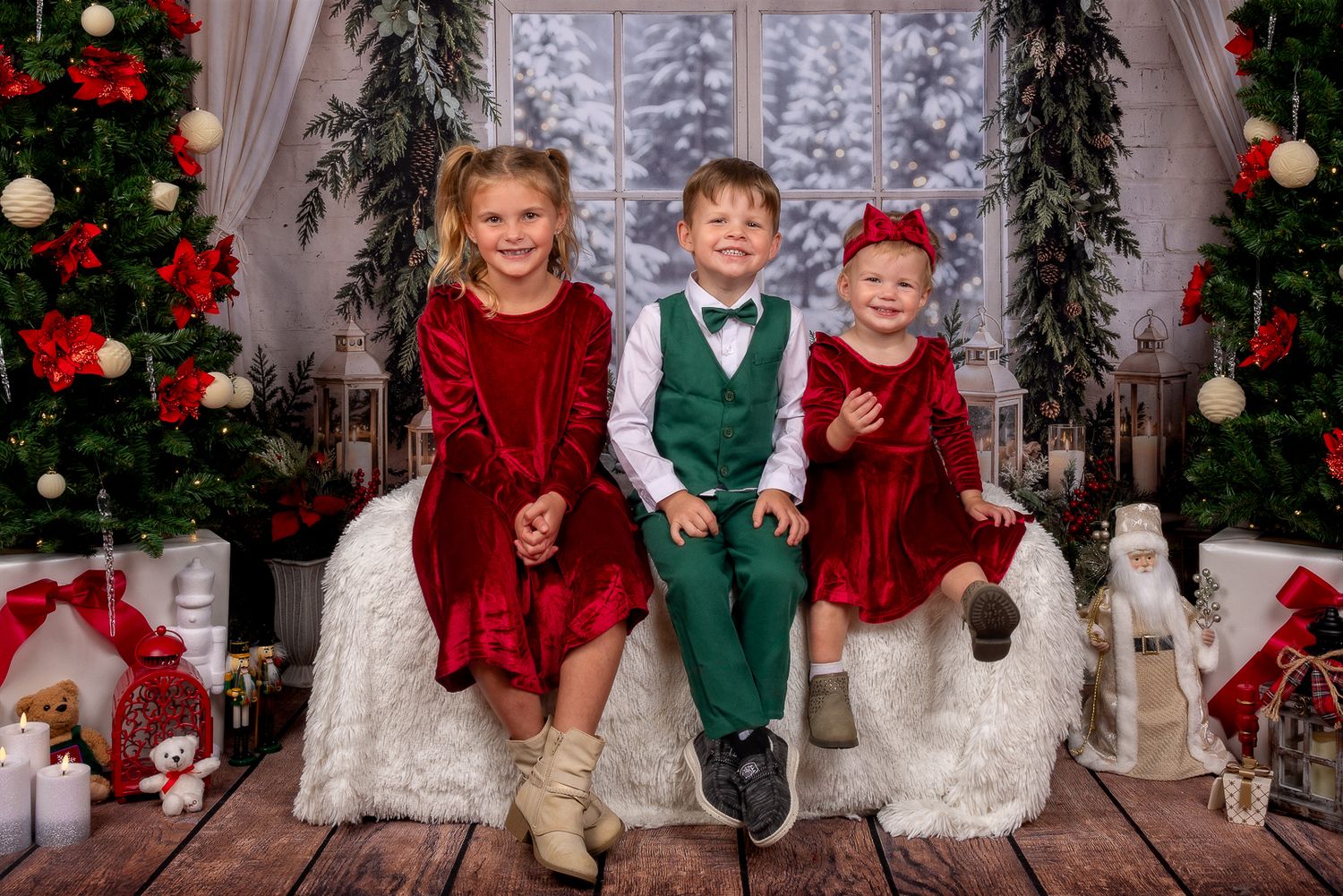 Three children in red velvet dresses and a green vest sit on a white fur rug in a festive Christmas mini session with trees and gifts.