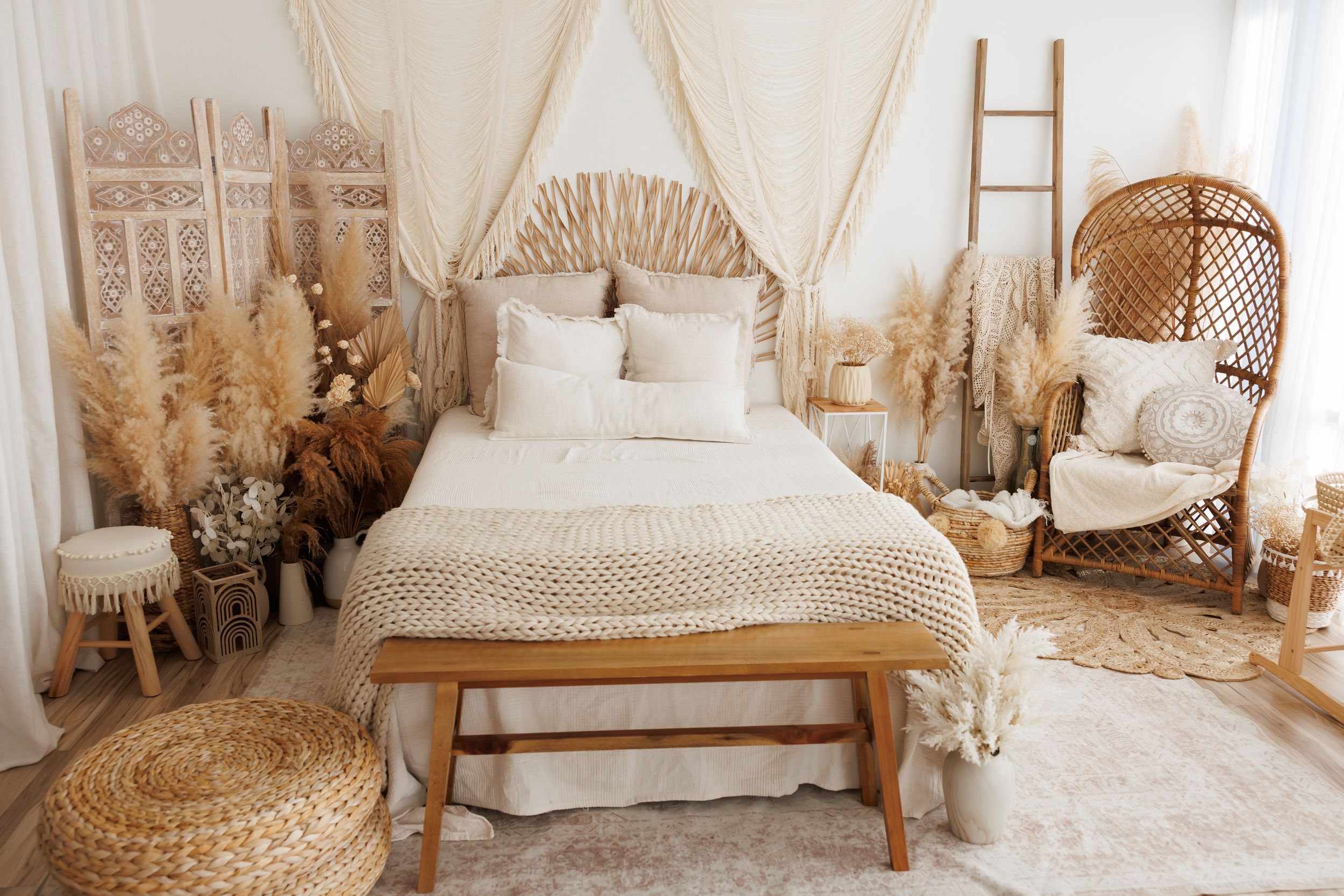 Cozy boho bedroom with a rattan headboard, neutral knit bedding, wooden bench, wicker peacock chair, and dried pampas grass decor.