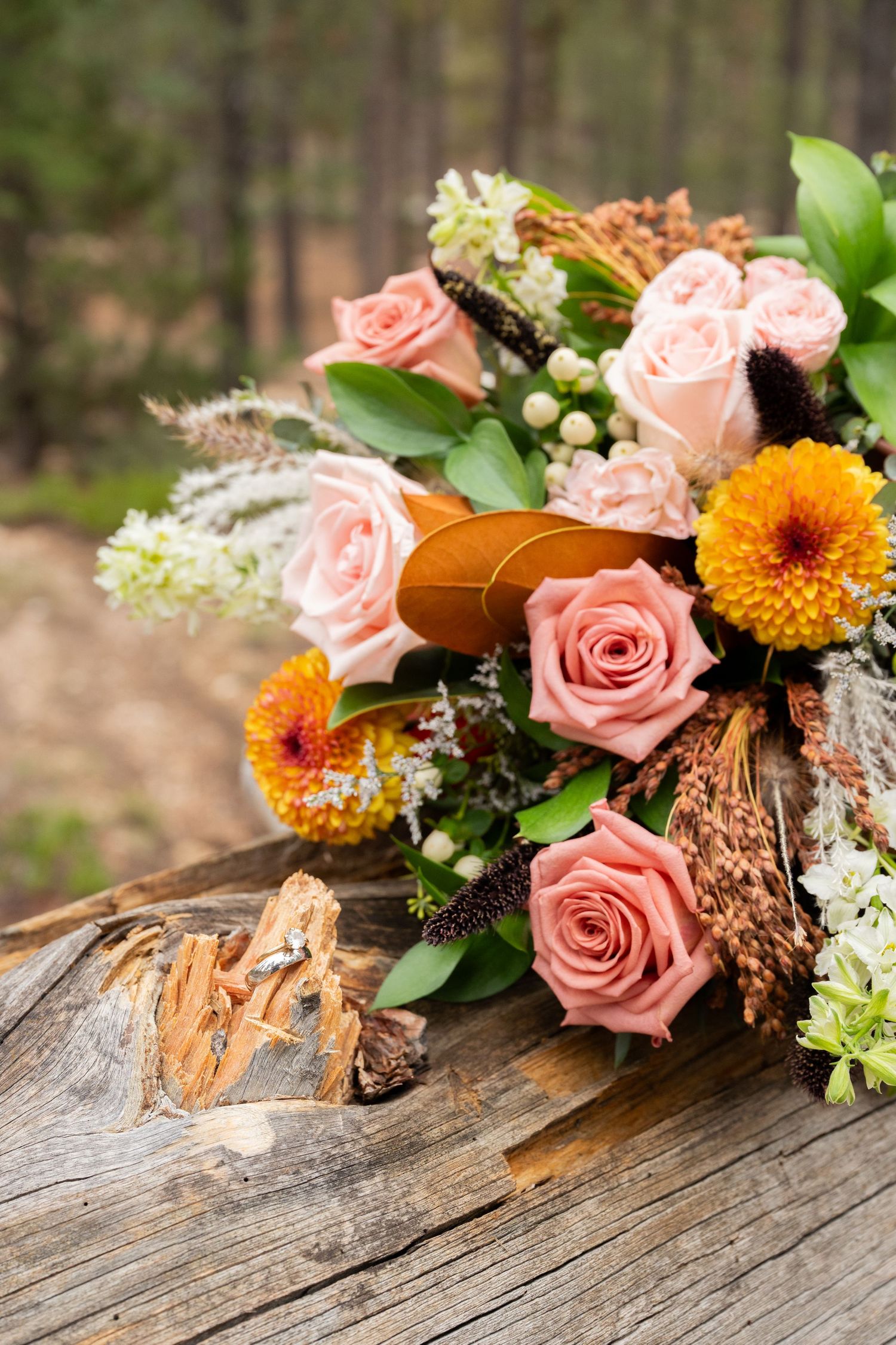 Rustic wedding bouquet with pink roses and orange flowers on weathered wood log in forest setting.