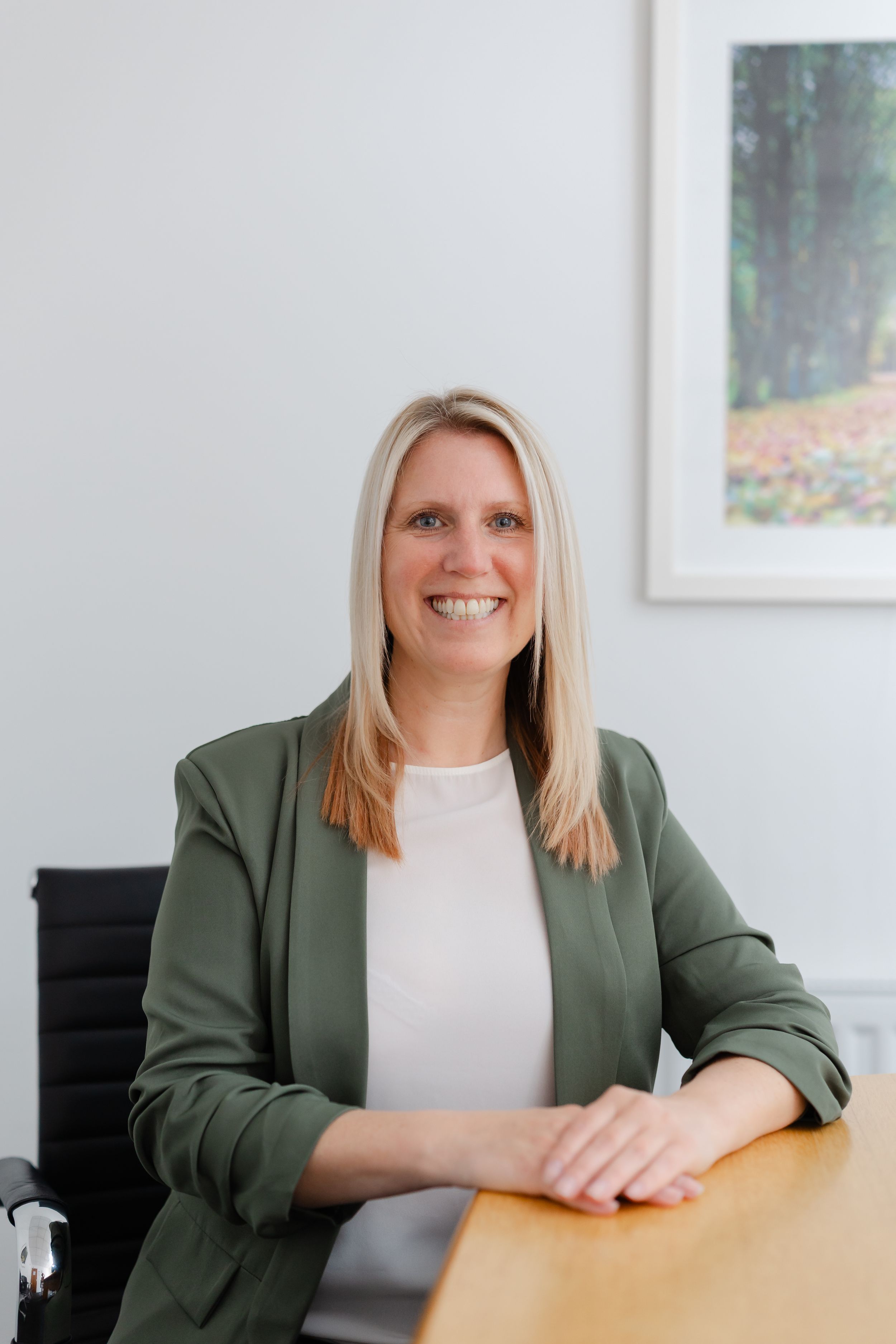 Professional woman in green blazer smiling at desk in modern office with artwork on wall.
