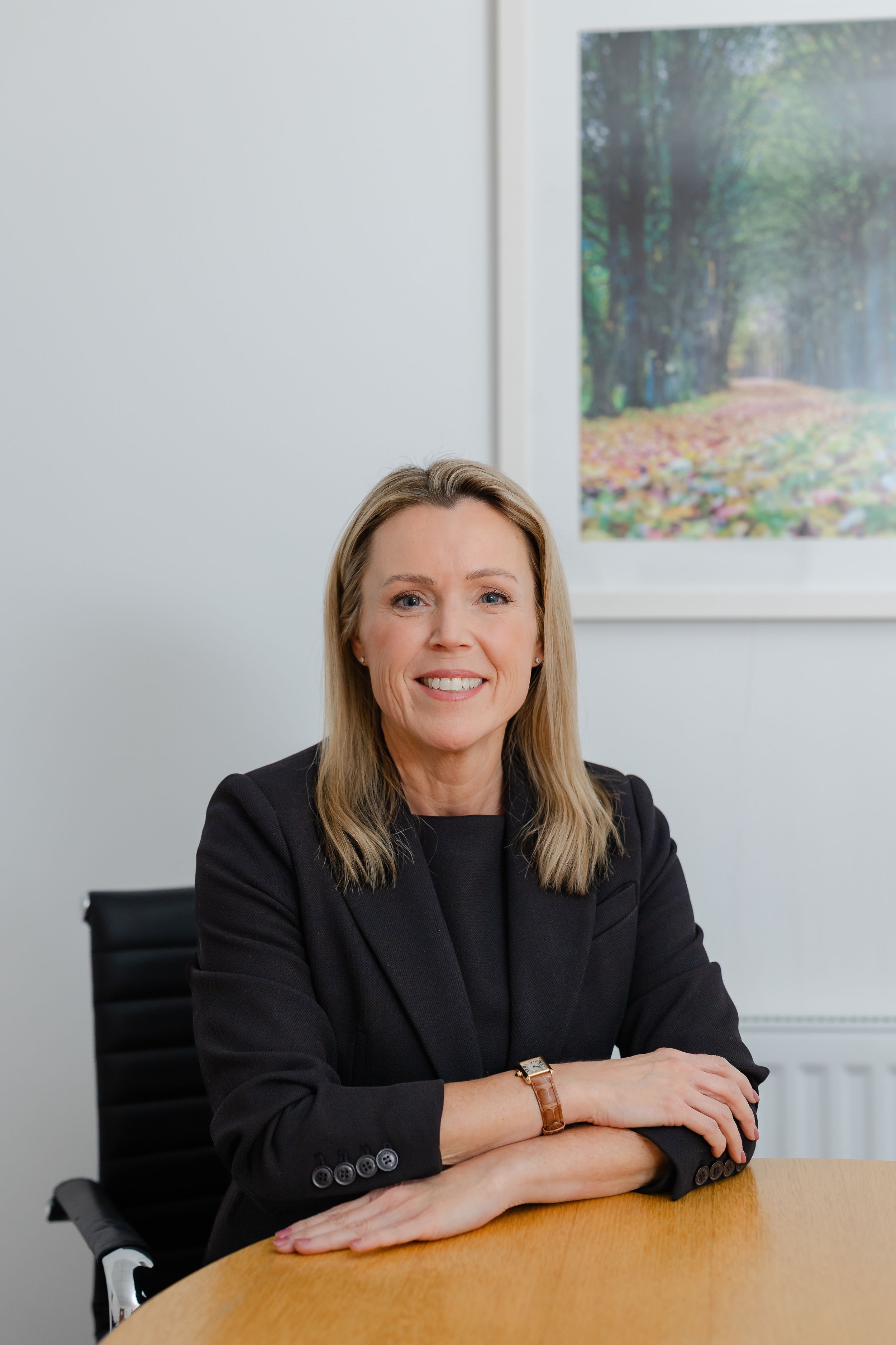 Professional woman in black blazer sitting at desk with arms crossed, smiling confidently in a modern office.