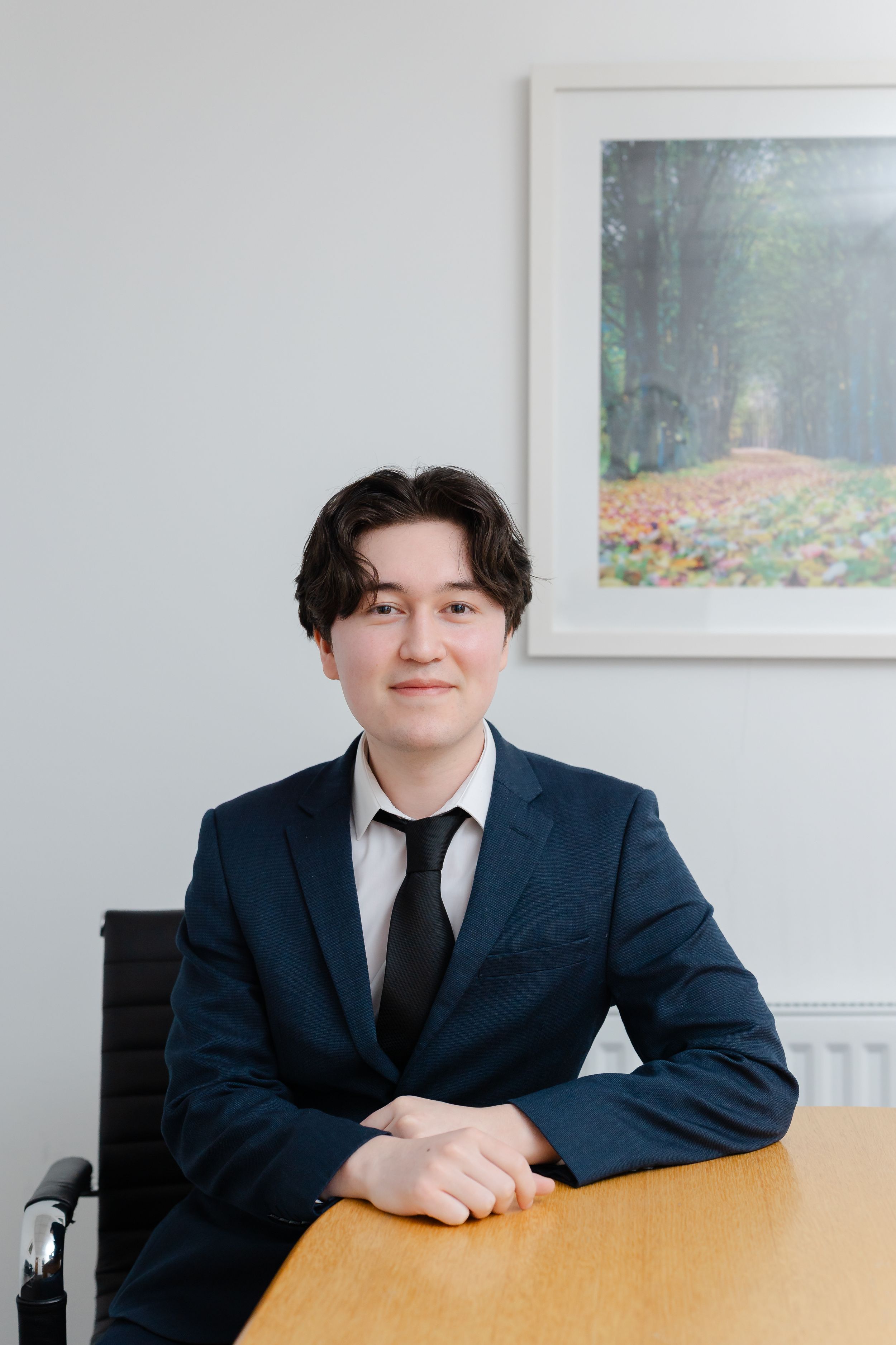 Young professional man in navy suit and tie seated at a wooden desk in a bright modern office setting.