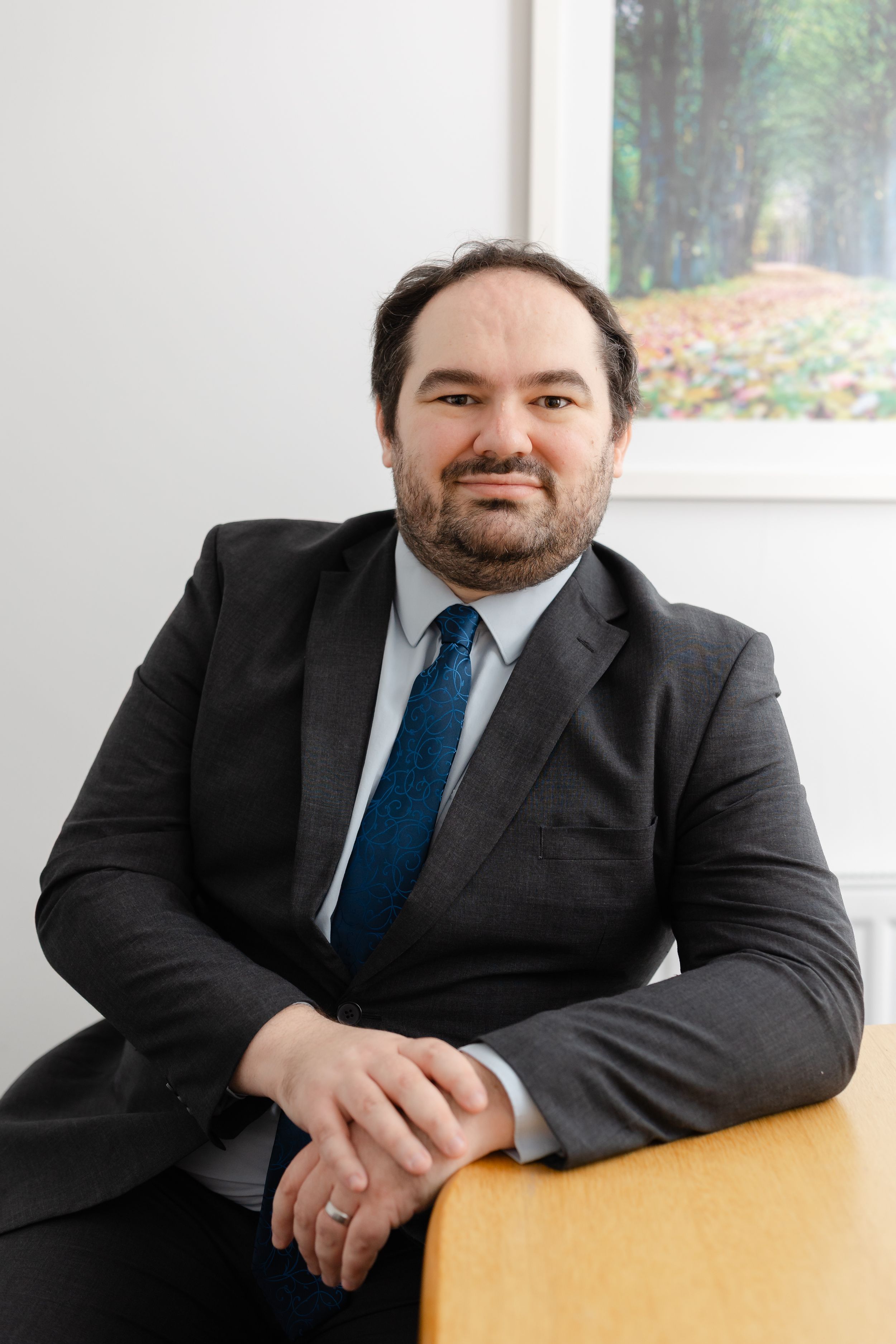Professional man in dark suit and blue tie sitting relaxed, smiling in a modern office setting.
