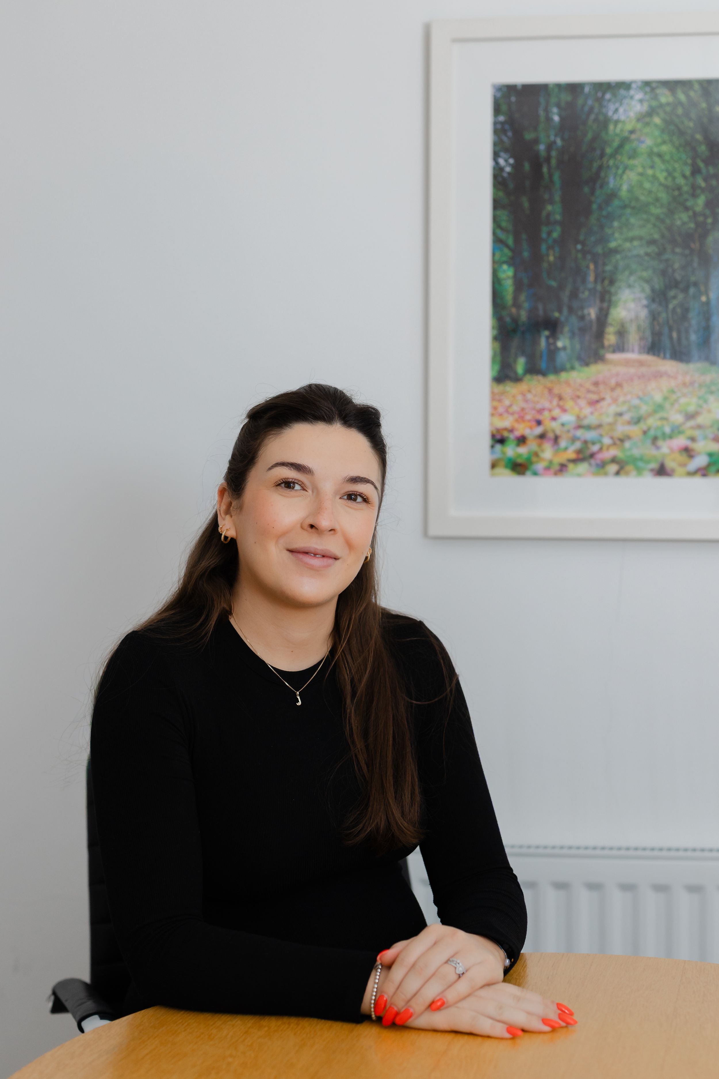 A young woman with long brown hair in a black top sits at a wooden desk, smiling, with a floral artwork behind her.