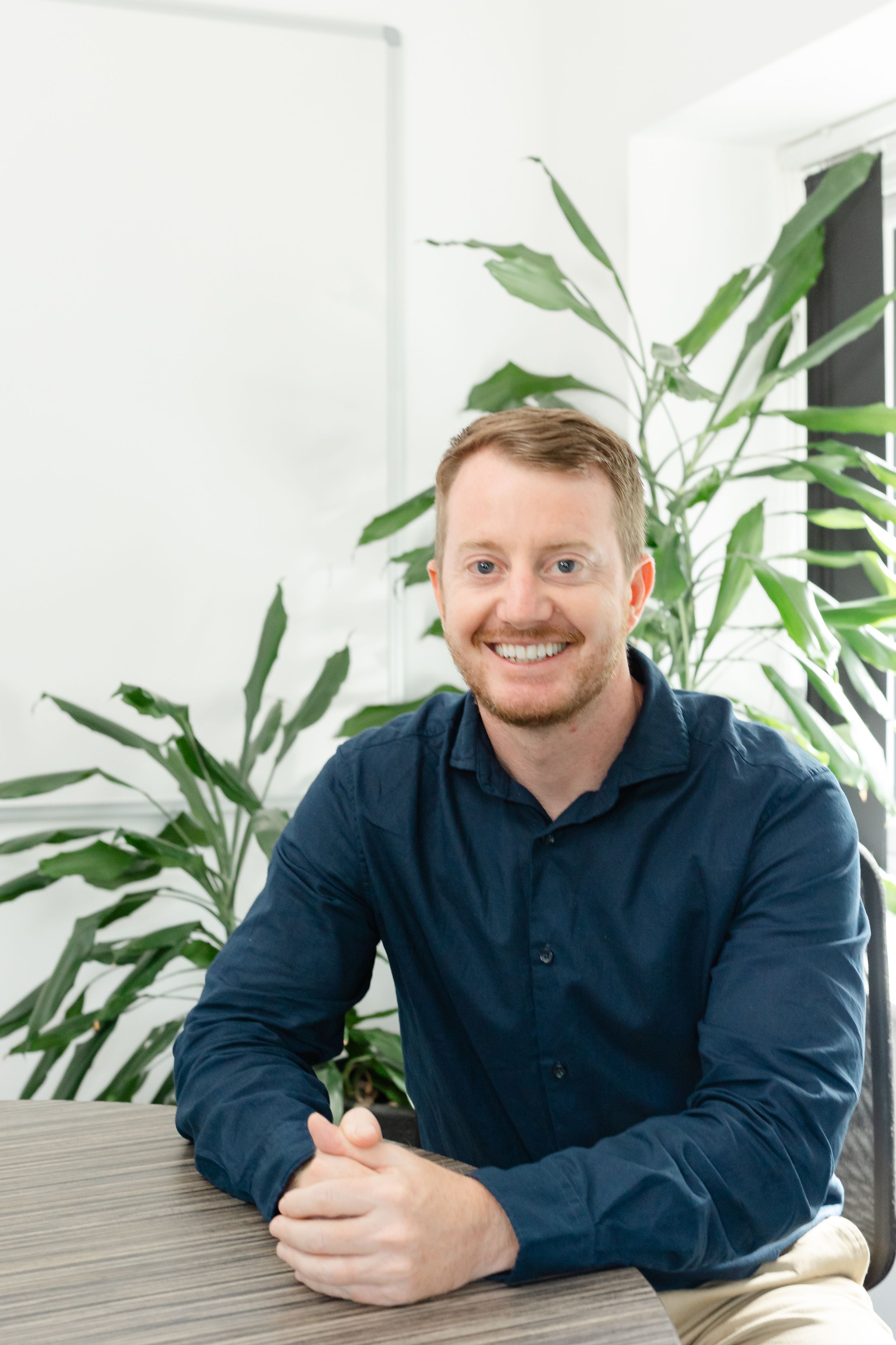Smiling young man with red hair in navy shirt sits at table with green plants in bright office.