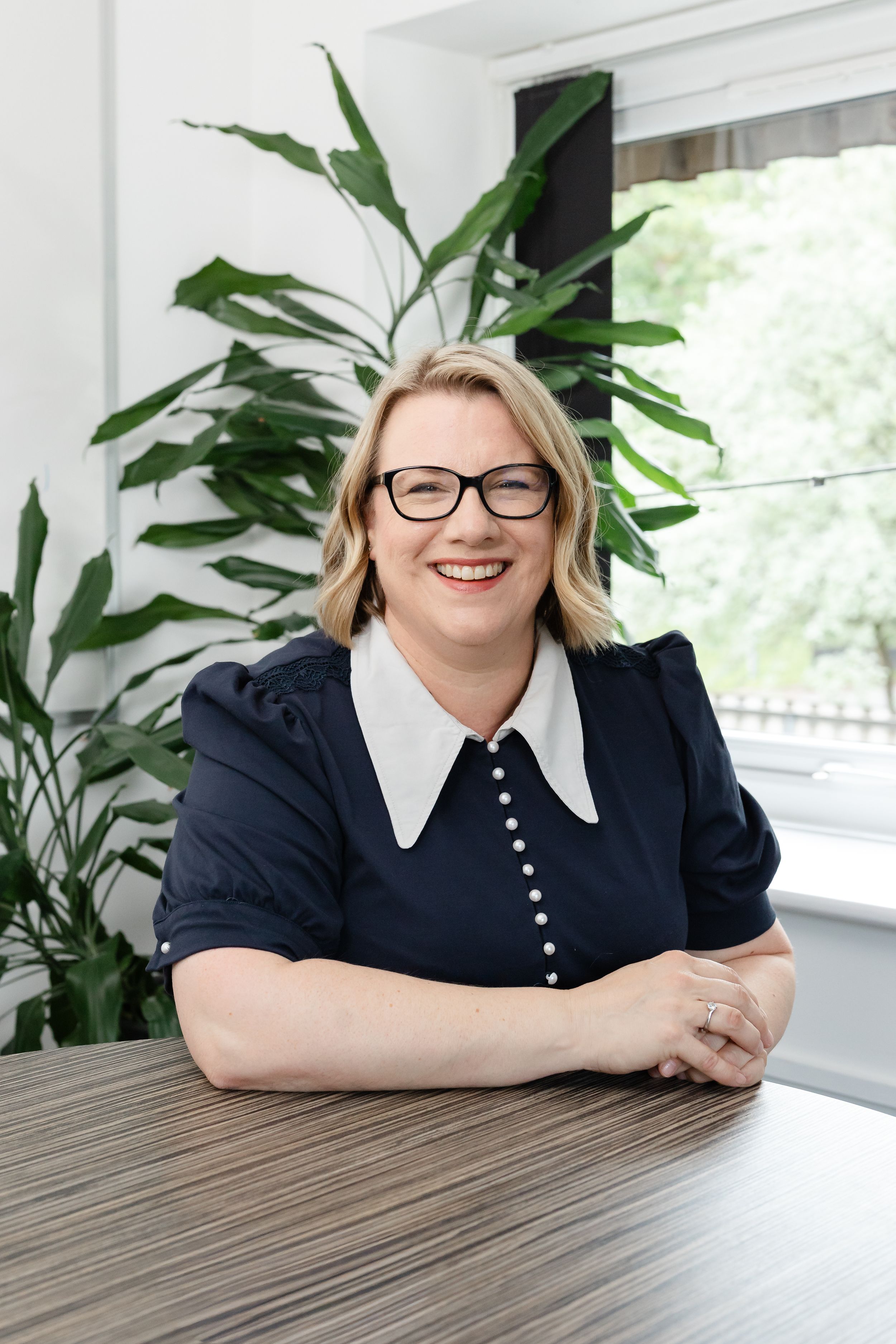 Smiling blonde woman with glasses in navy dress sits at wooden table with green tropical plant in bright office.