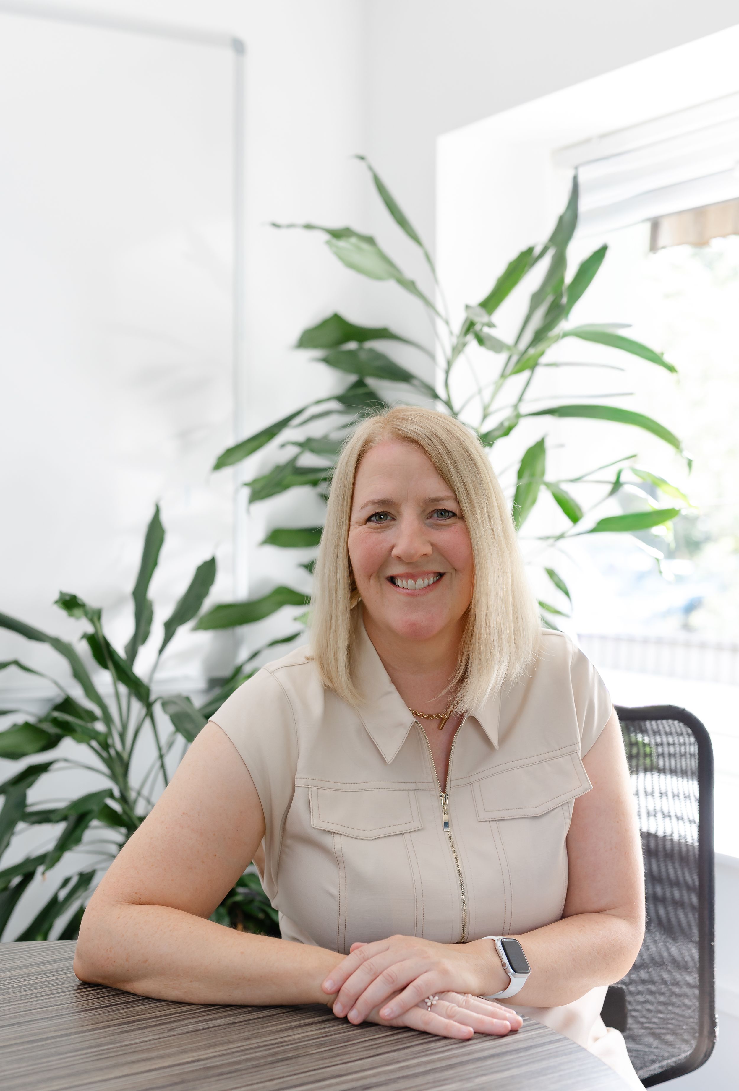 Smiling blonde woman in beige sleeveless top seated at a table with green tropical plants in a bright modern office.