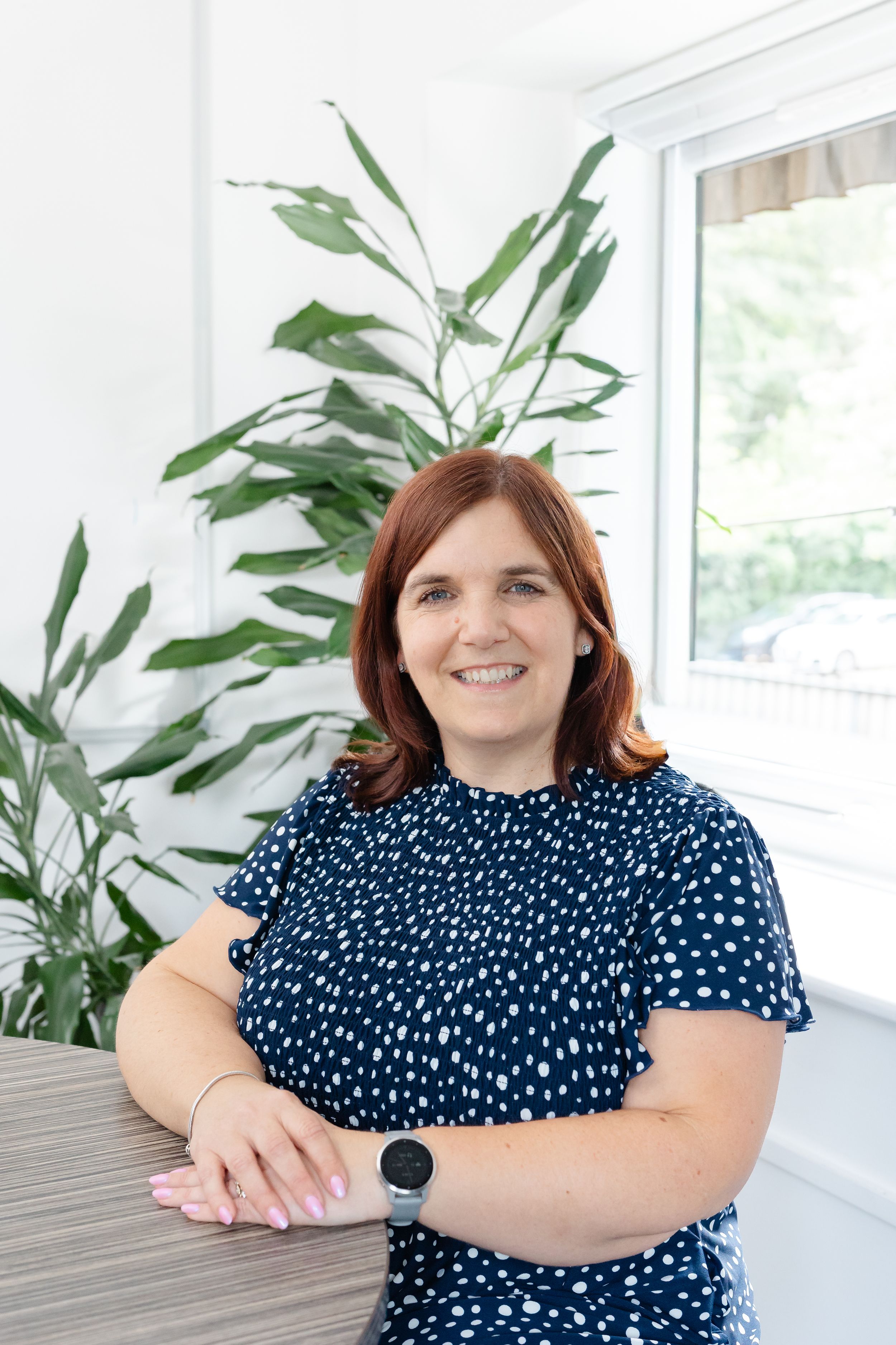 Smiling woman in navy polka dot dress seated by a table with green plants and window in bright office.
