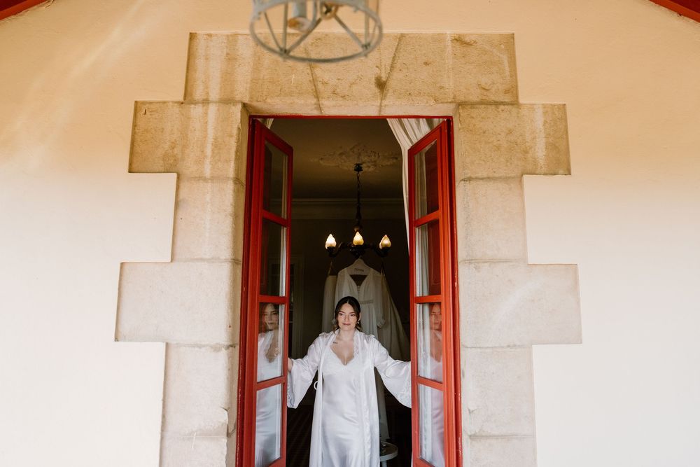 Bride in a white robe stands in an open doorway with red wooden doors, framed by a stone archway, with a chandelier glowing behind her.