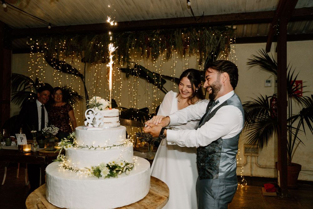 Smiling bride and groom cut their tiered white wedding cake together at a rustic reception with string lights and a sparkler.