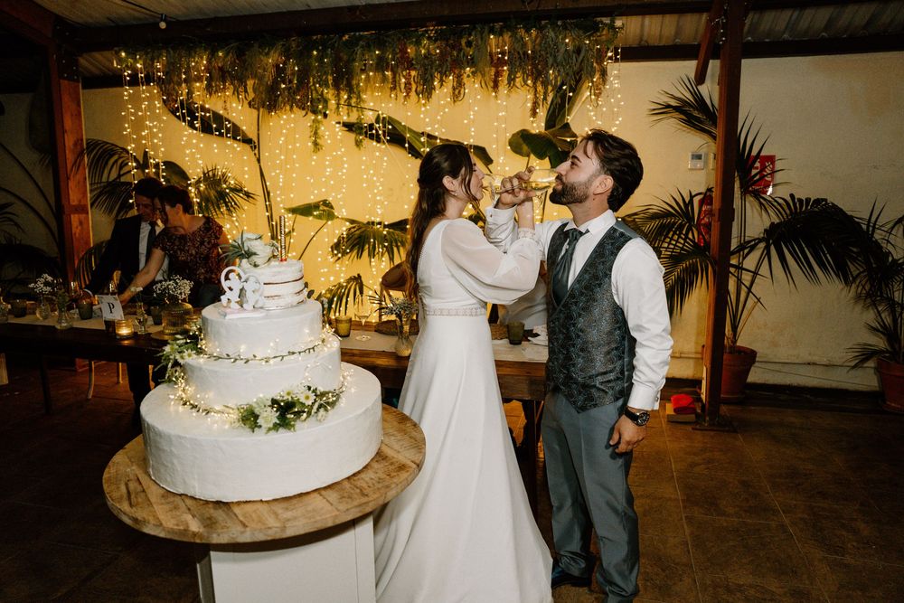 Bride in a flowing white gown and groom in a vest share a toast beside a tiered wedding cake at a rustic reception with string lights.