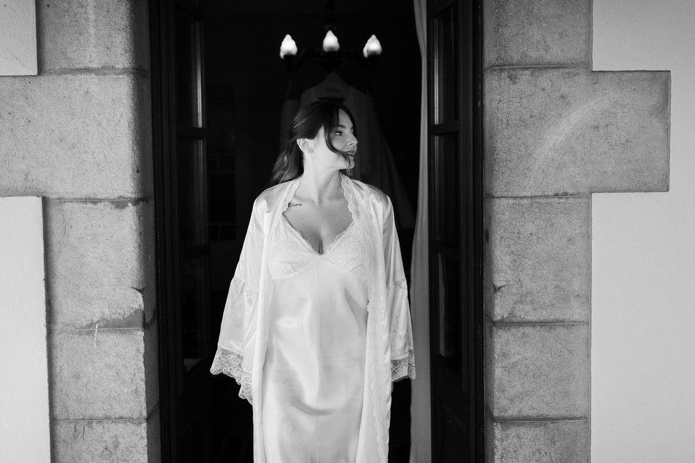 Black and white photo of a woman in a white silk robe and slip dress standing in a stone doorway, glancing over her shoulder.