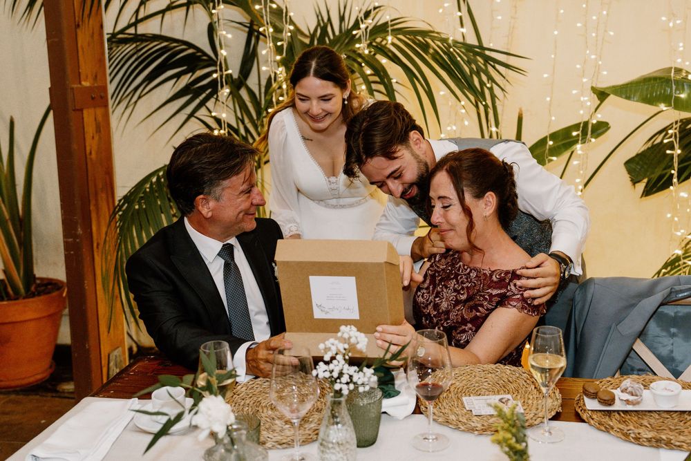 Smiling woman in a white dress and a man lean over a seated couple at a festive table, sharing a cardboard gift box at a celebration.