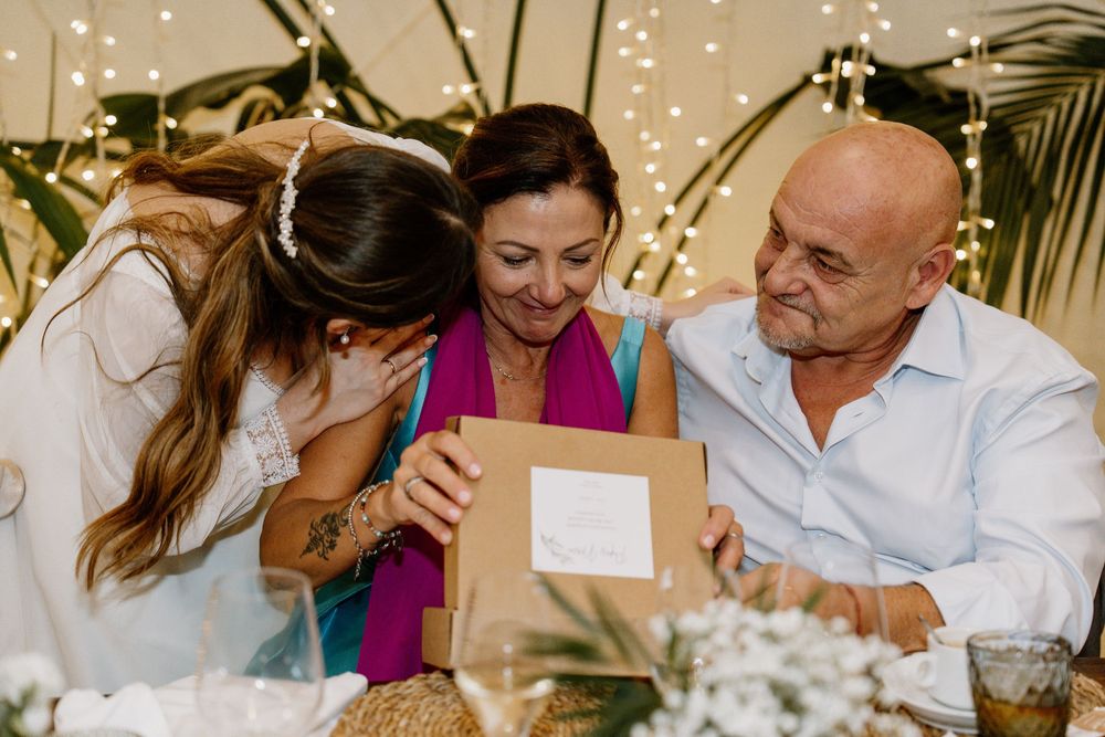 Bride hugs a smiling woman holding a gift box as an older man watches, capturing a heartfelt moment at a wedding reception table.